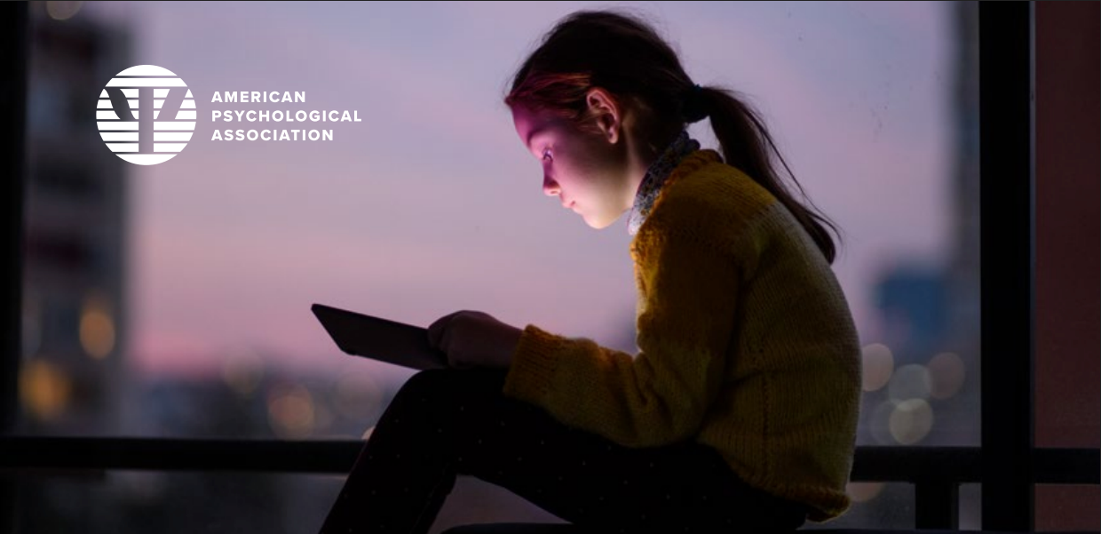 A young girl sitting by a window at dusk, looking at a tablet device, with city lights in the background. The image features the American Psychological Association logo.