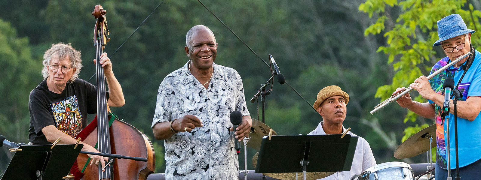 Four musicians performing outdoors, one playing a double bass, one singing into a microphone, one on drums, and one playing a flute, with a green leafy background.