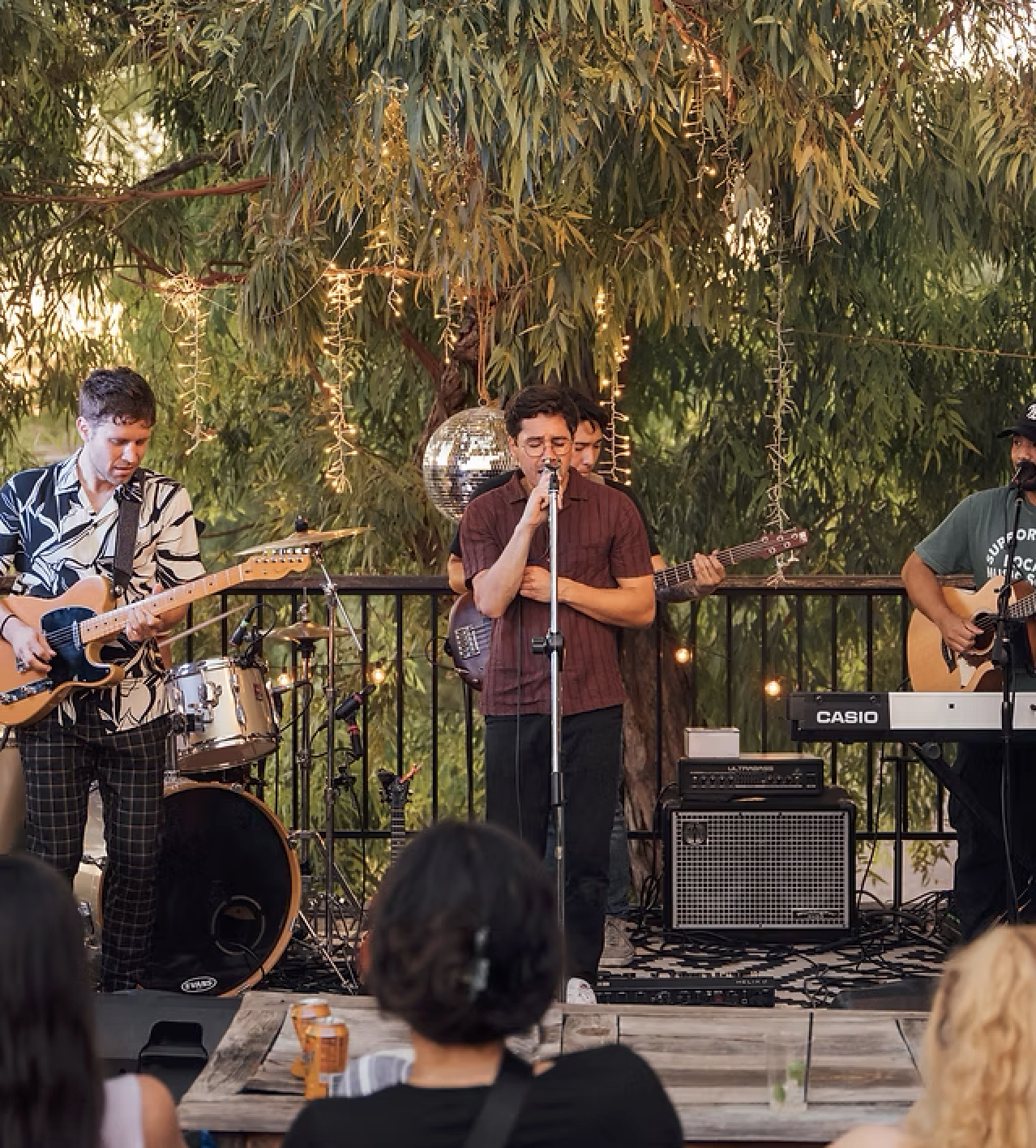 Four musicians perform on an outdoor stage decorated with string lights and a disco ball, with audience members watching.