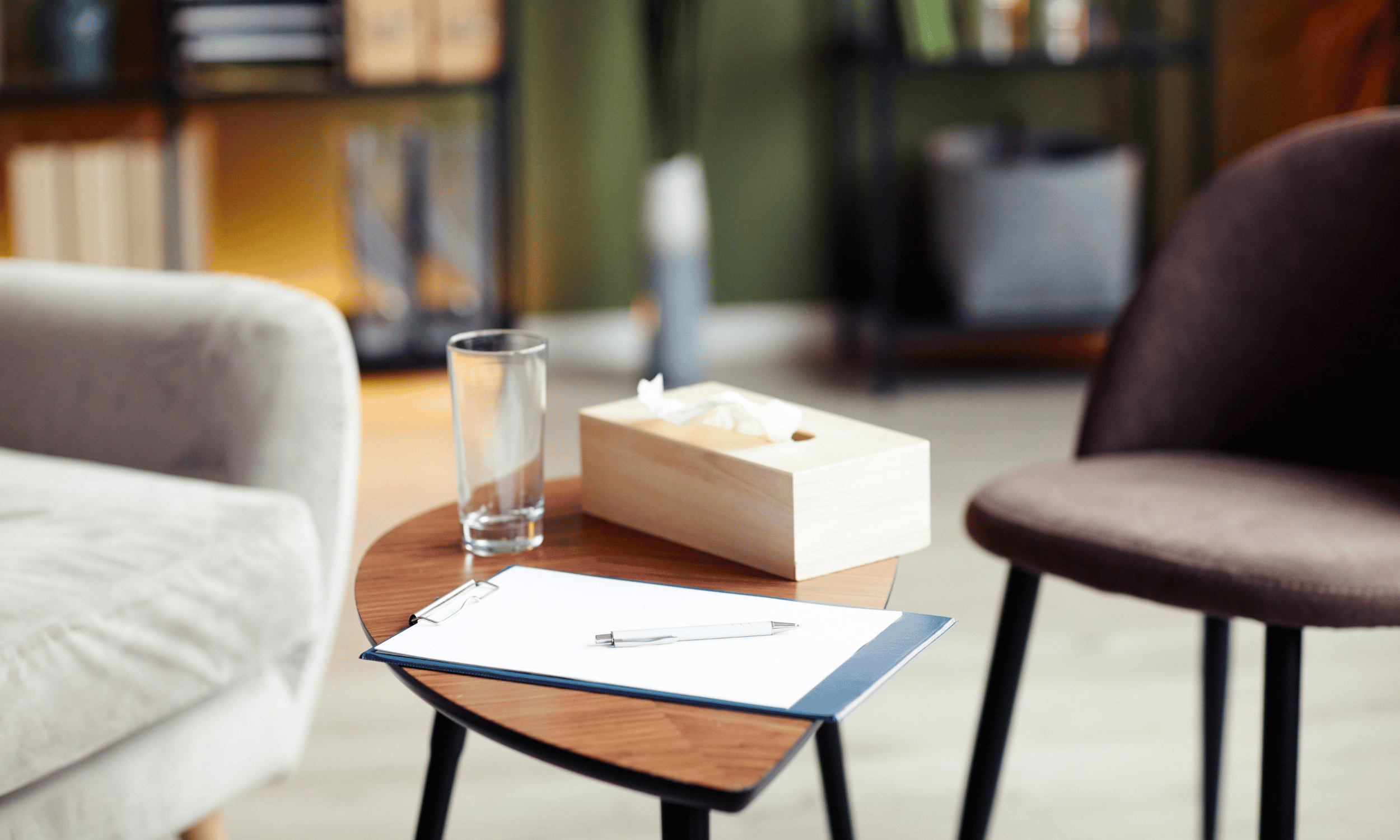 Close-up of a wooden table with a glass of water, a tissue box, a clipboard with paper and pen inside, and two chairs, one cream-colored and one dark brown, in a room with bookshelves and a green wall in the background.