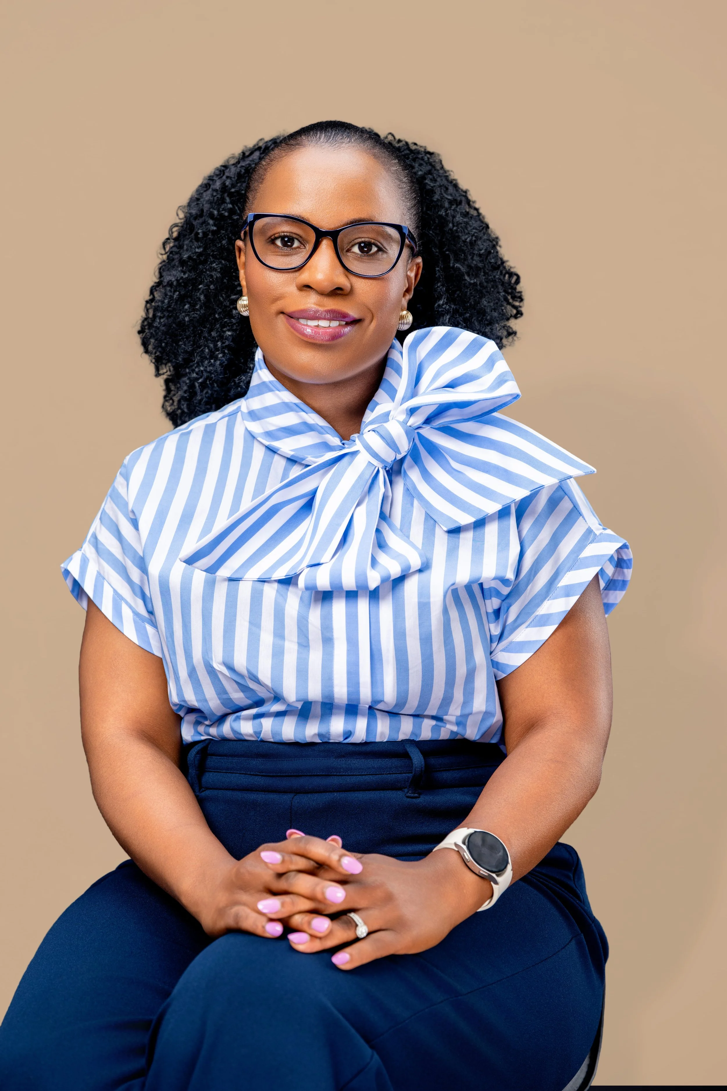 Portrait of a woman with curly black hair wearing glasses, a blue and white striped blouse with a bow, blue pants, and a white watch, sitting against a beige background.