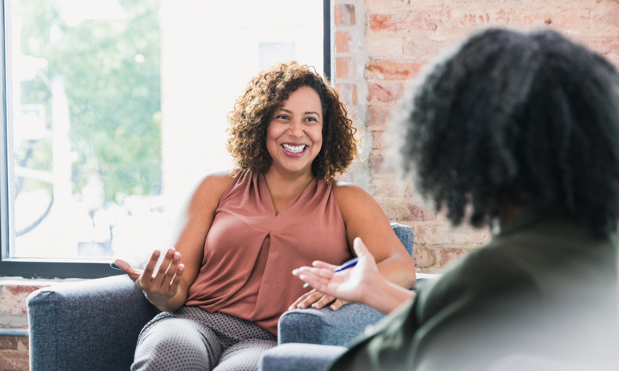 A woman with curly hair smiling and talking to someone across from her in a casual indoor setting with exposed brick wall and large window.