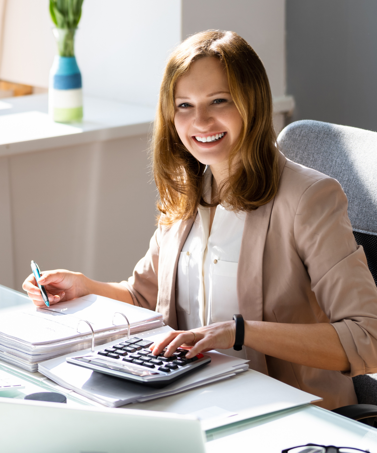 A smiling woman with red hair sitting at an office desk, holding a pen in one hand and using a calculator with the other.