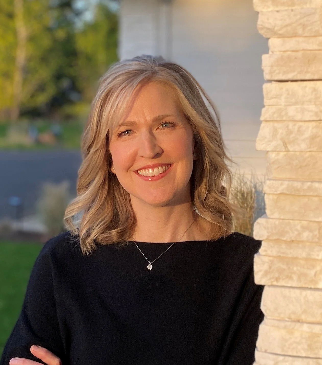 A smiling woman with blonde, shoulder-length hair wearing a black top and a silver necklace, standing near a beige stone wall outdoors during sunset.