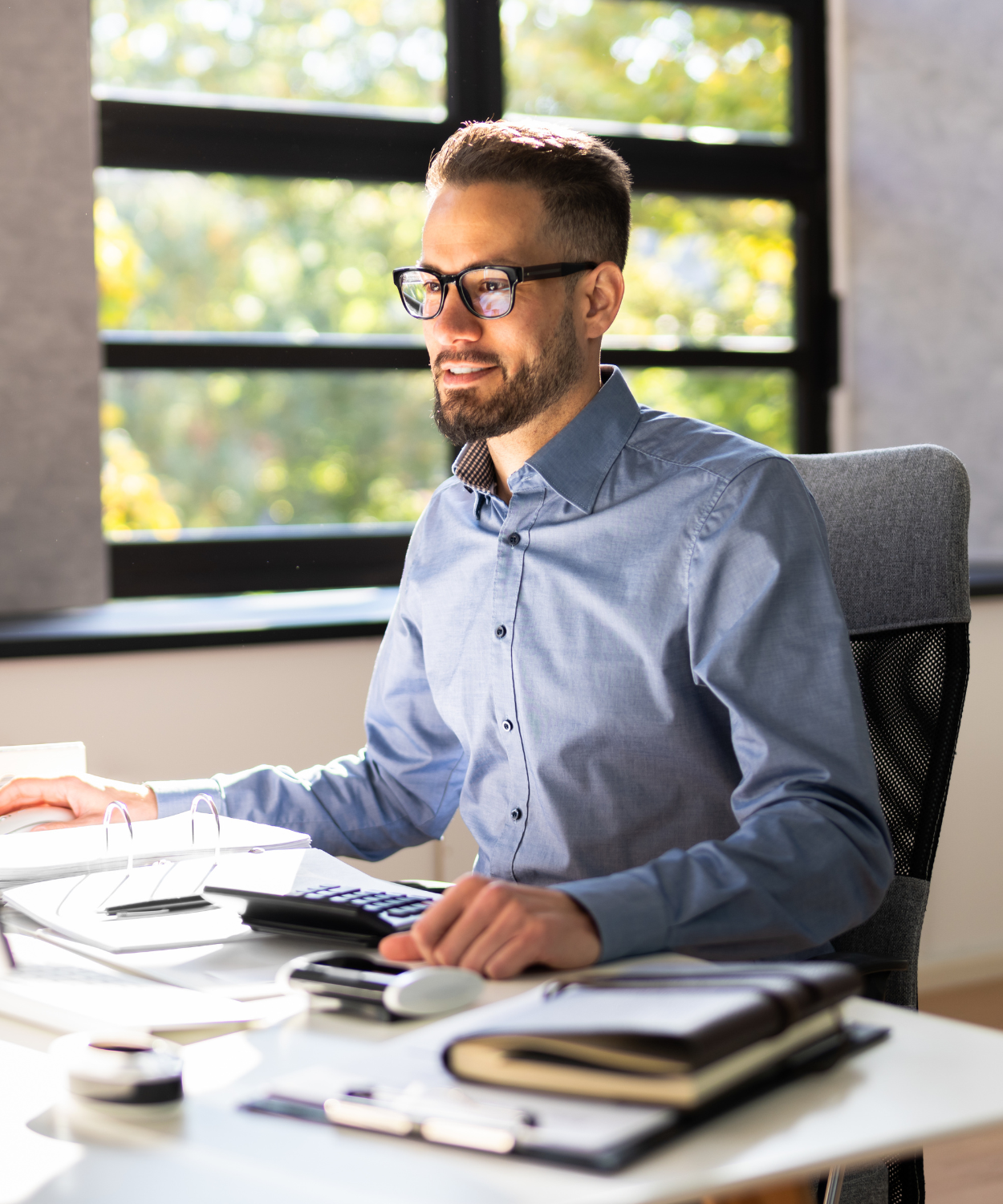 A man with glasses and a beard sitting at a desk in an office, working on a computer, with large windows showing trees in the background.