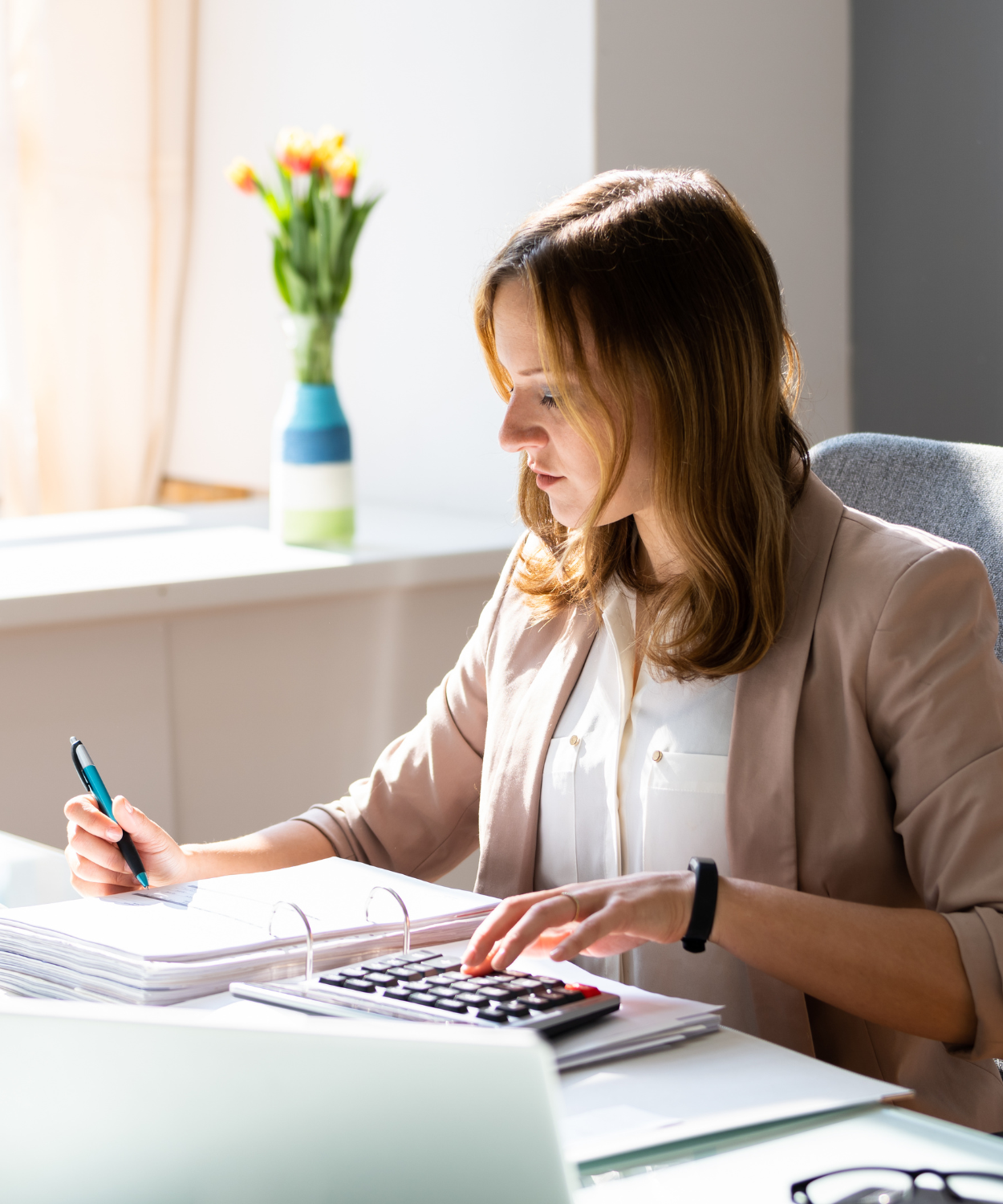 A woman working at a desk with a calculator, papers, and a pen, in a bright office with a vase of flowers in the background.