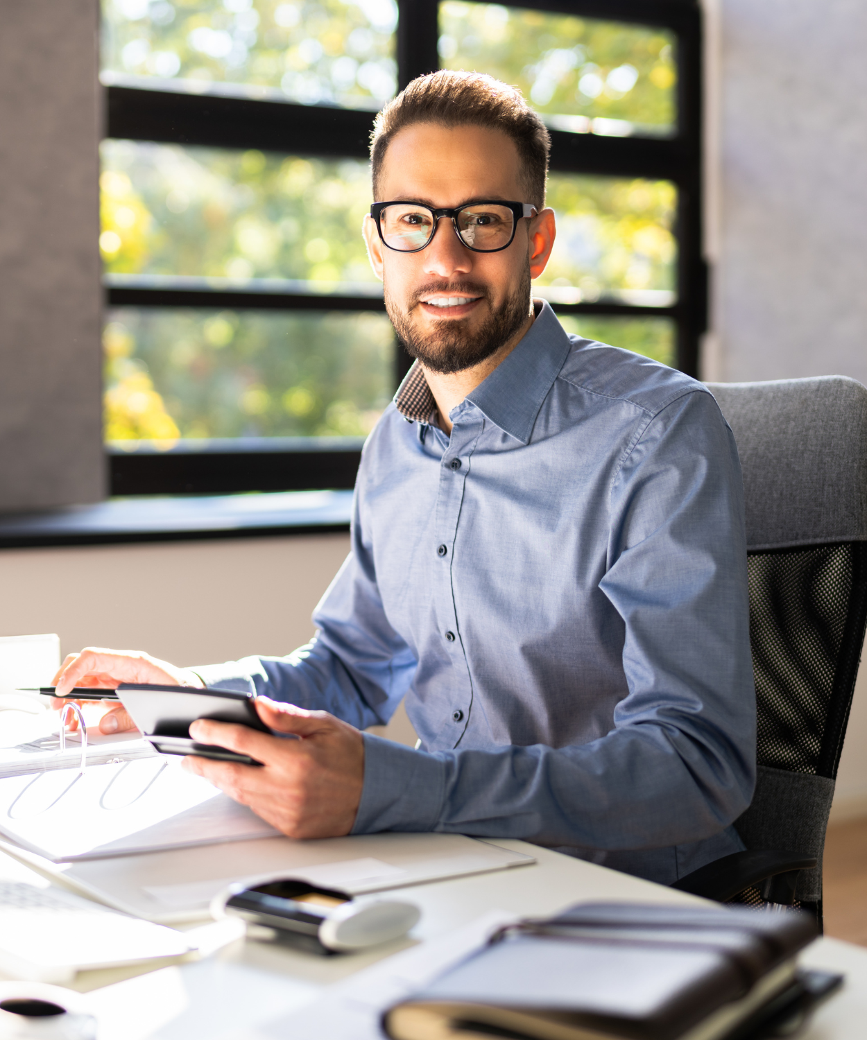 A young man with glasses and a beard sitting at an office desk, holding a smartphone, smiling, with books and papers on the desk, in front of a window with sunlight and trees outside.
