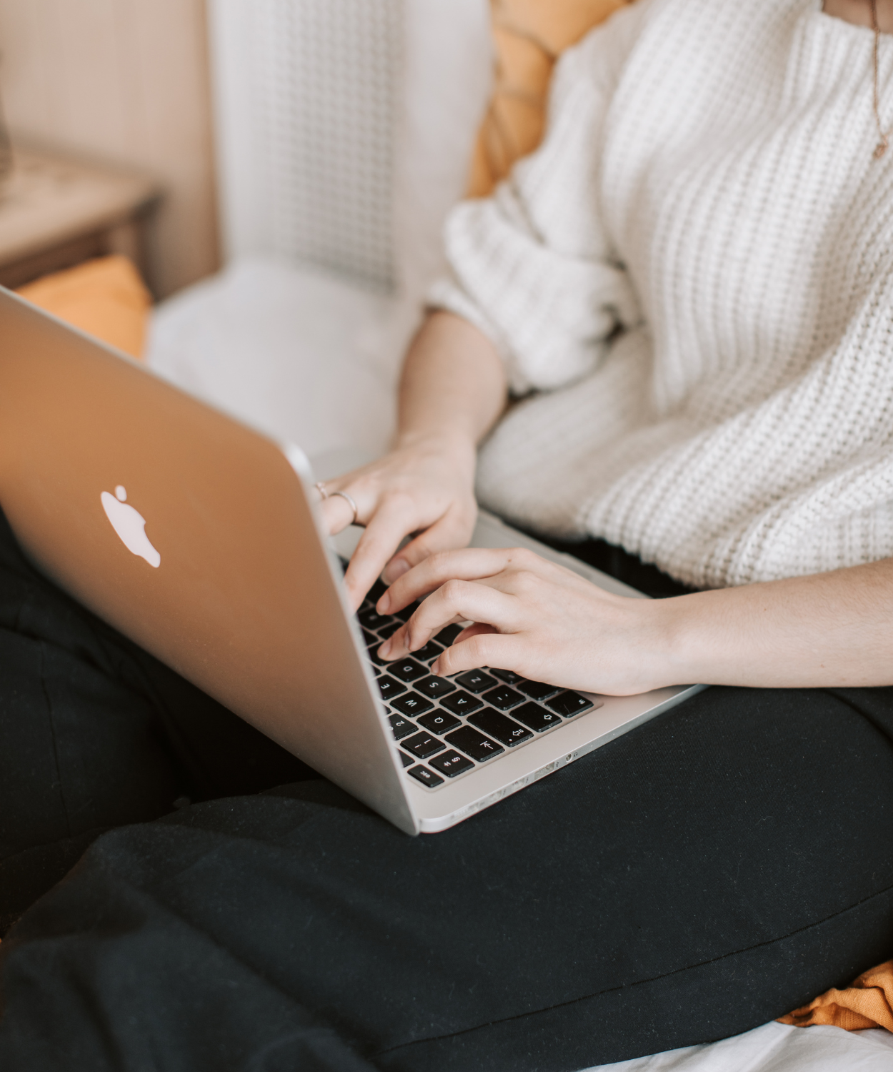 Person using a silver MacBook laptop, seated on a bed with beige bedding and a black blanket.
