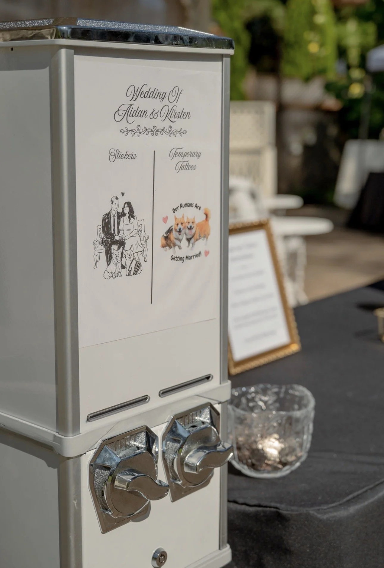 A white wedding sticker machine with signs indicating it offers stickers and temporary tattoos, featuring illustrations of a couple and dogs, at an outdoor wedding event.