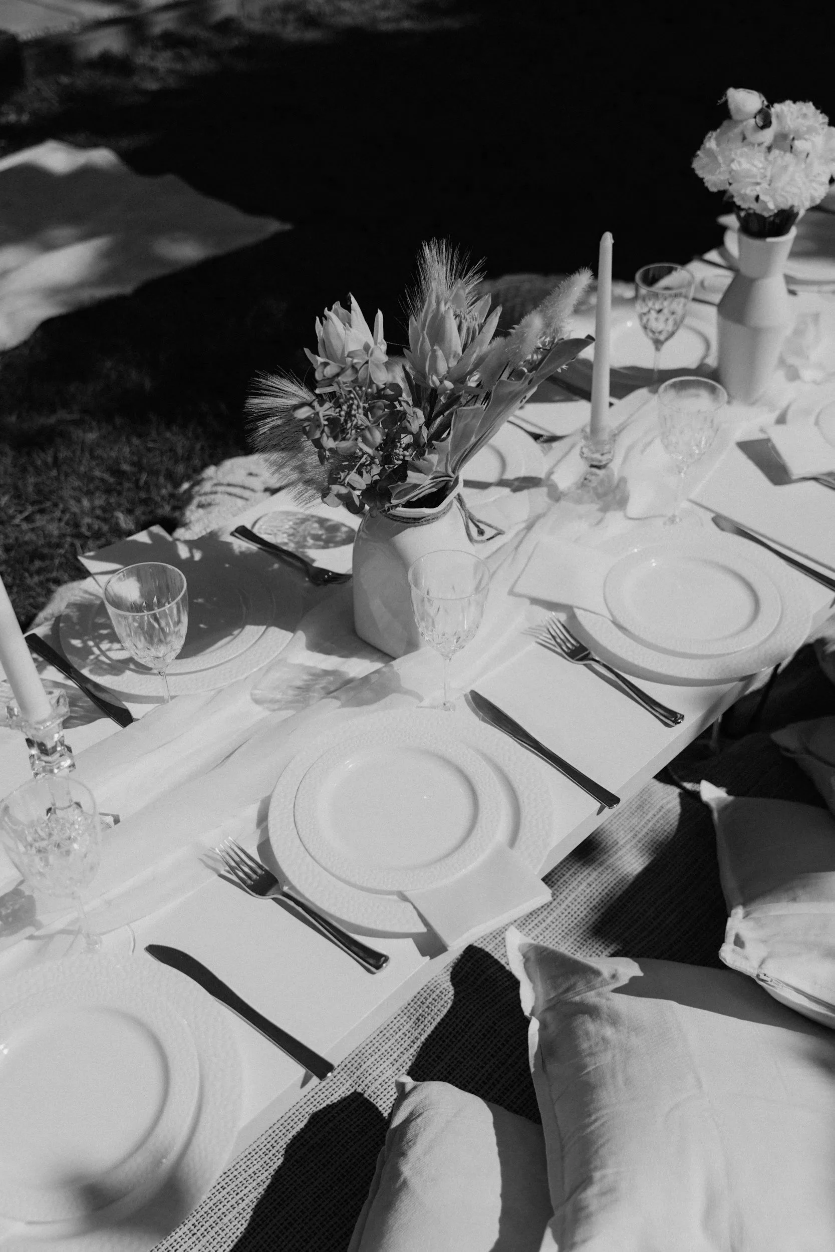 A black and white photo of an outdoor dinner table set with plates, glasses, cutlery, candles, and floral centerpieces.