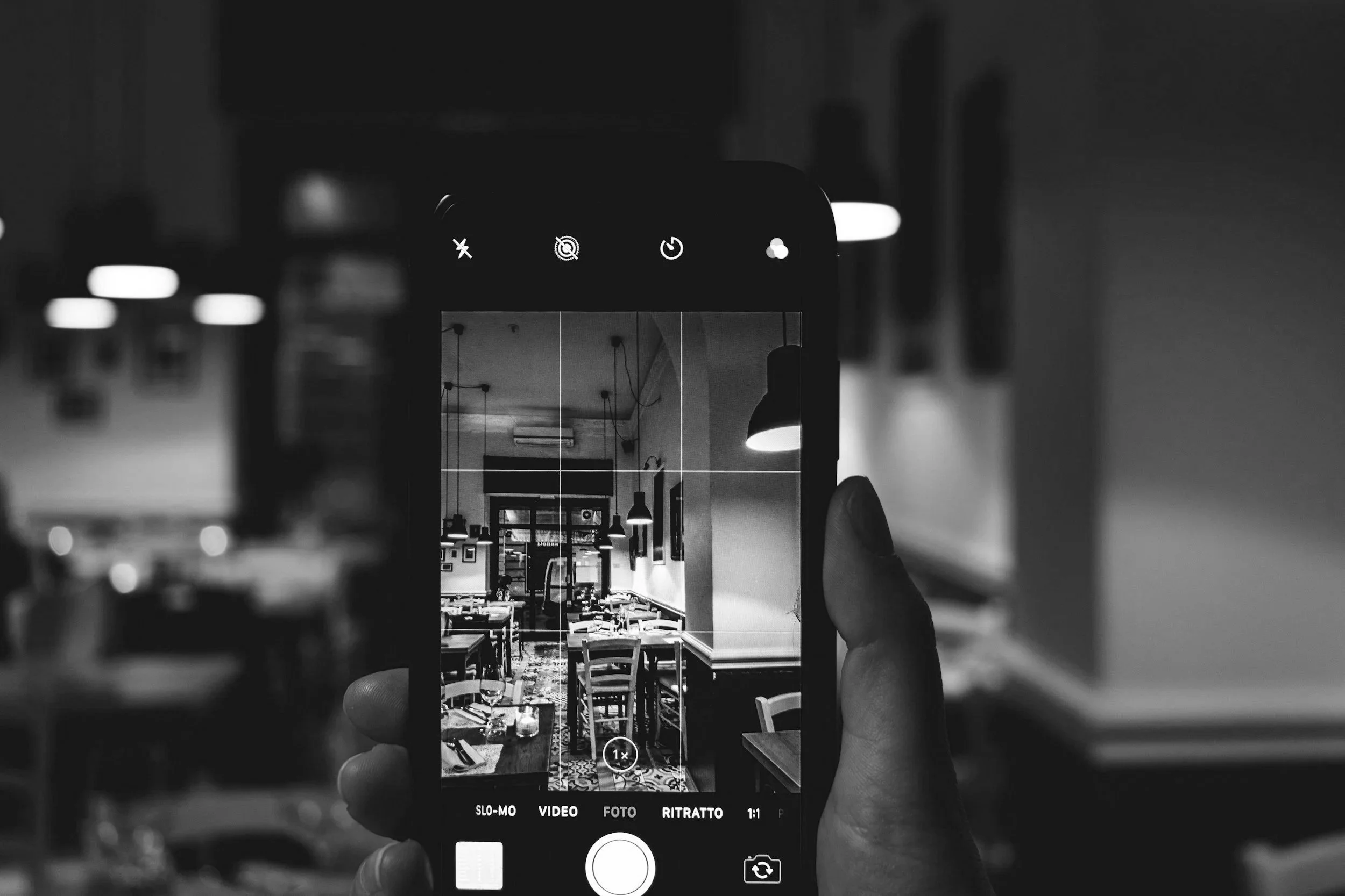A black and white photo of a person taking a picture of a restaurant interior through a smartphone's camera interface. The restaurant has tables, chairs, pendant lights, and framed pictures on the walls.
