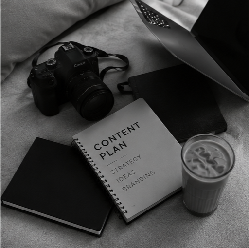 A camera, a laptop, a notebook, a content plan notebook, and a glass of iced coffee on a desk.