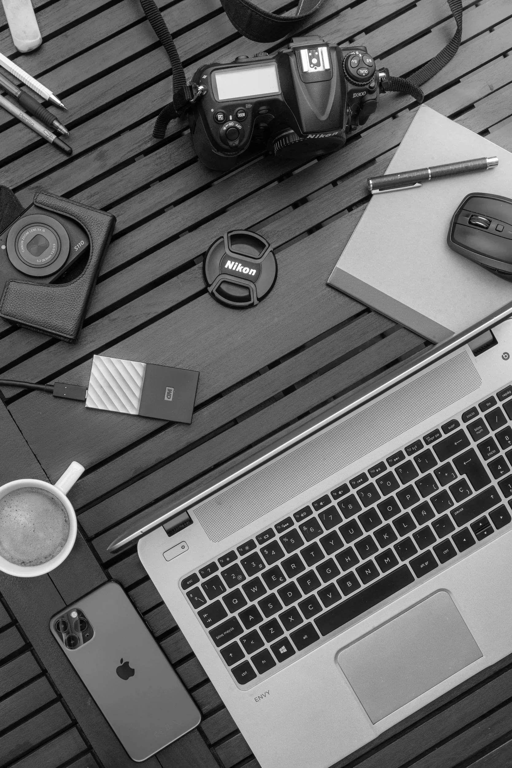 Black and white digital camera, Nikon lens cap, portable hard drive, Apple iPhone, notebook, wireless computer mouse, notebook, laptop, printed document, coffee mug, pen, phone lens cap, backpack strap on wooden surface with a cup of coffee.
