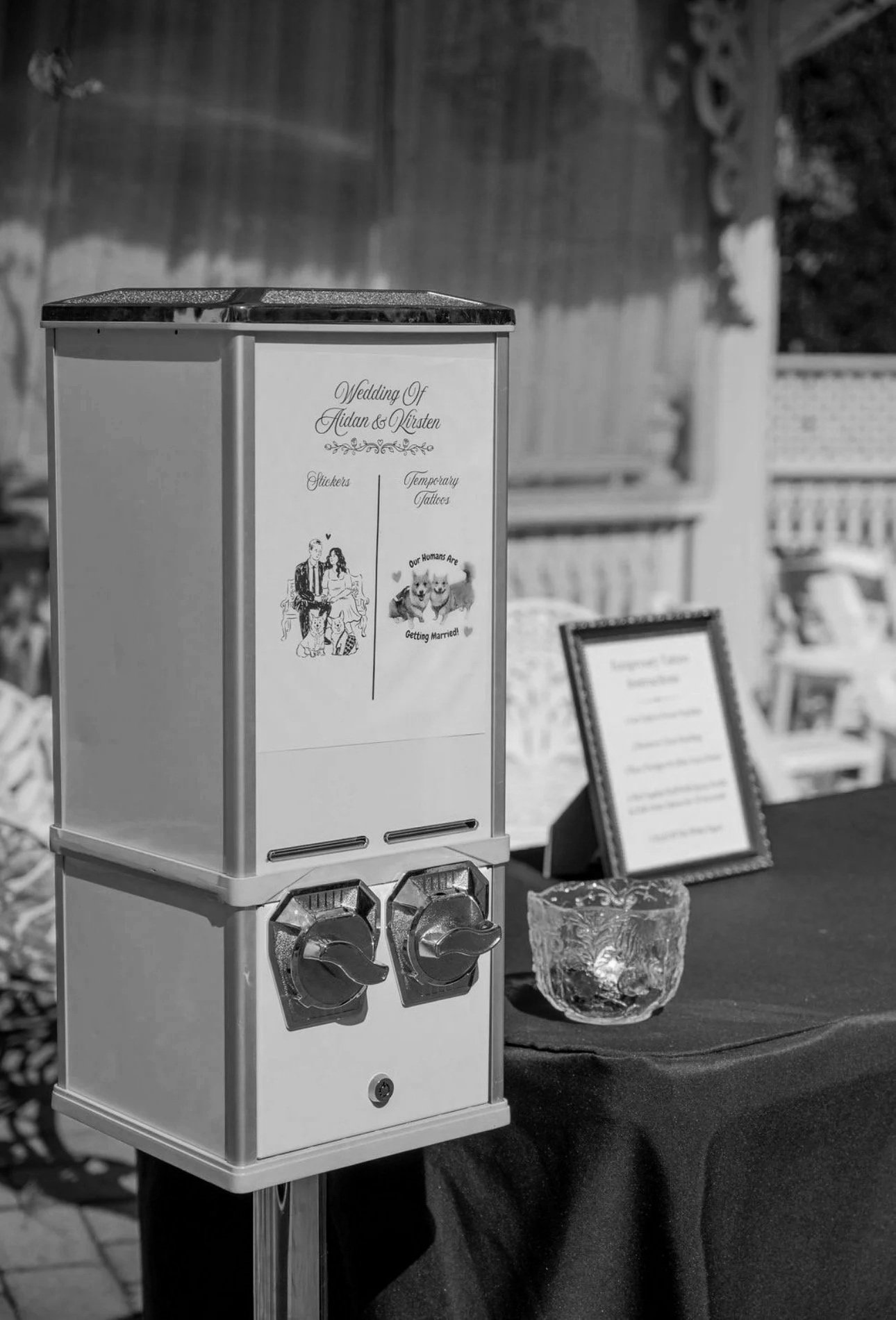 A sticker machine on display at a wedding reception with signs indicating wedding details and fun facts. The background shows part of a decorated outdoor venue.