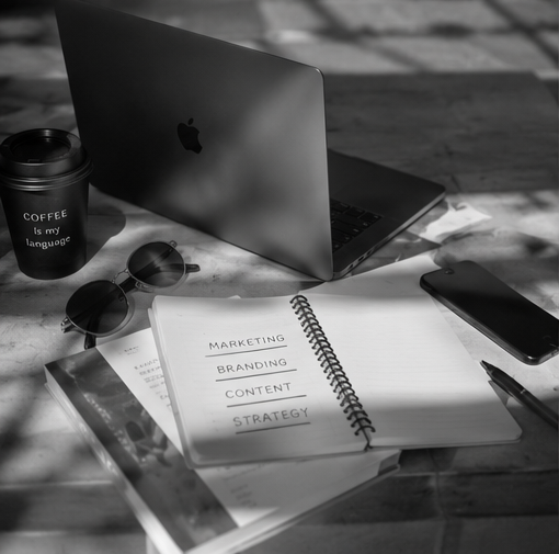 A desk with an open laptop, a coffee cup with the text "COFFEE is my language," a pair of sunglasses, a smartphone, and a notebook with words "Marketing, Branding, Content, Strategy" on the open page in black and white.