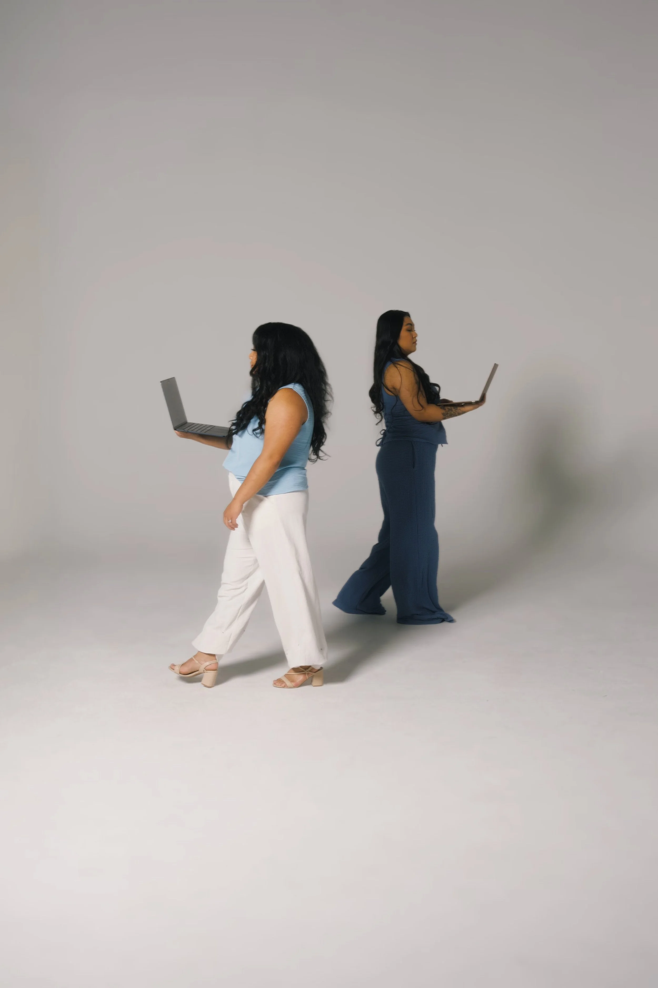 Two women standing against a gray wall, each holding a laptop and looking at their screens.