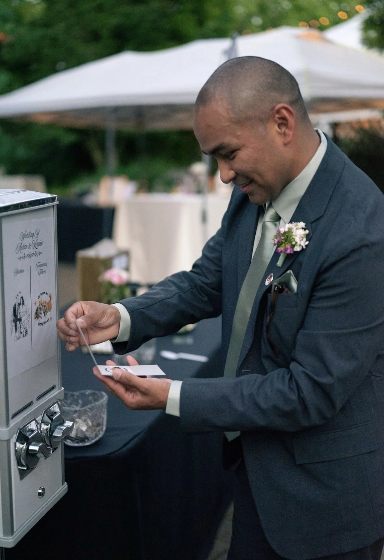 A man in a dark suit with a boutonniere is smiling and getting a sticker from a sticker machine at an outdoor event with a tent and string lights in the background.