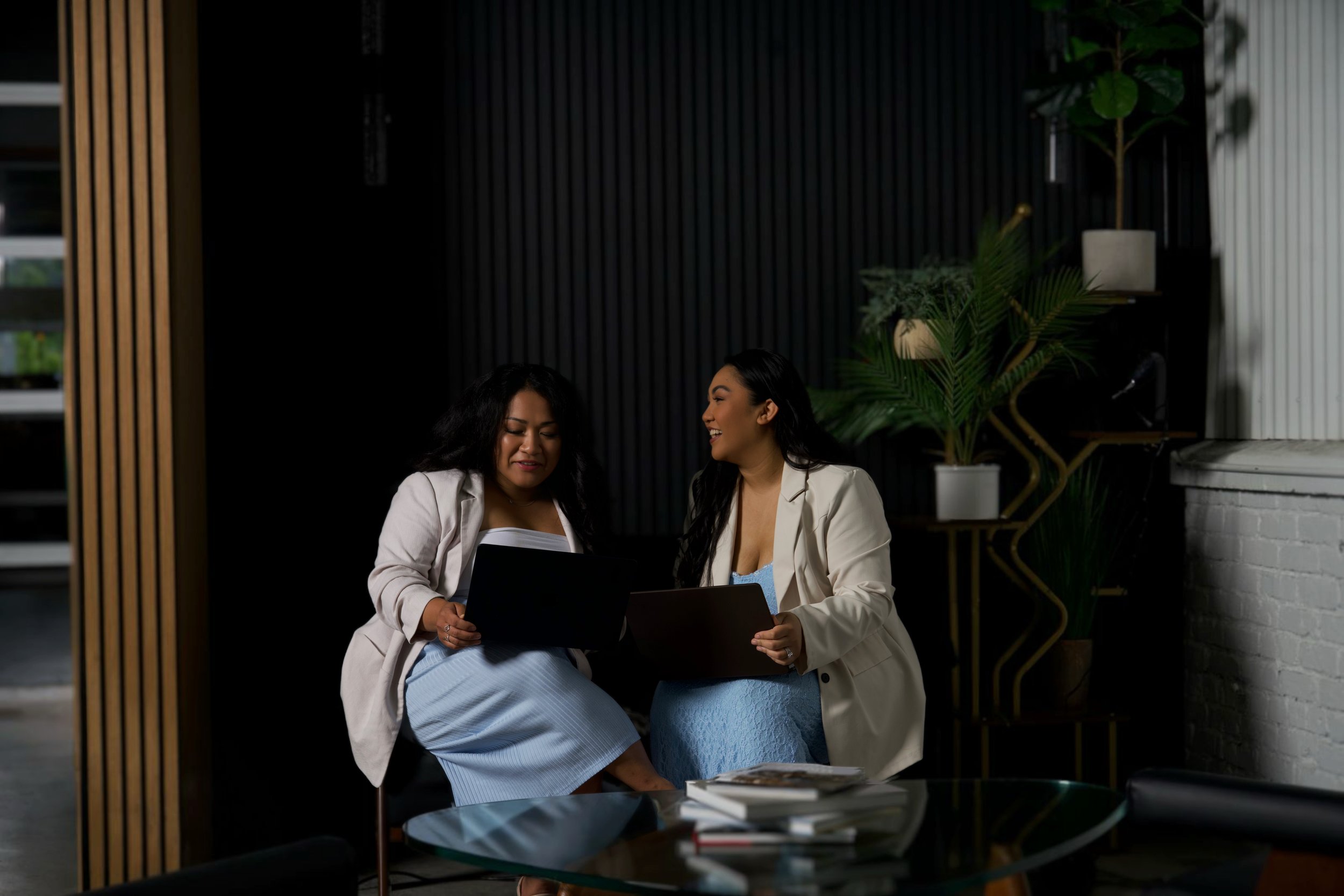 Three women sitting at a wooden table in a cozy coffee shop, engaged in conversation. The table has a laptop, a notebook, a cup of coffee, and a green drink. The shop has large windows, indoor plants, framed artwork, and a sign that reads 'Kynd Kreative'.