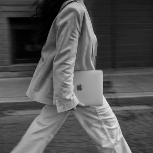 Person wearing a light-colored suit walking outside while holding a closed MacBook laptop.