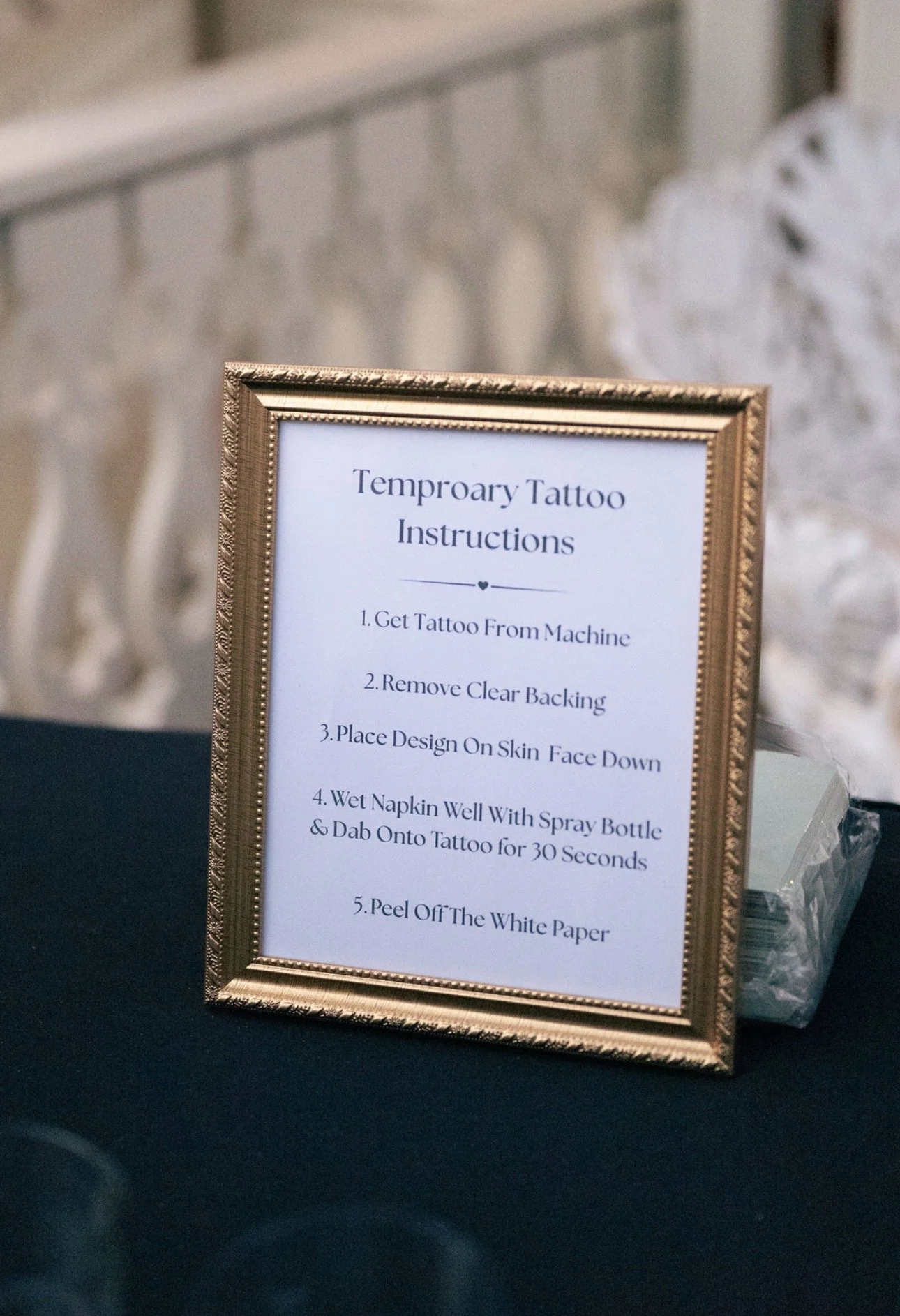 Sign with tattooing instructions, framed in gold, on a black surface, with a stone or brick wall in the background.