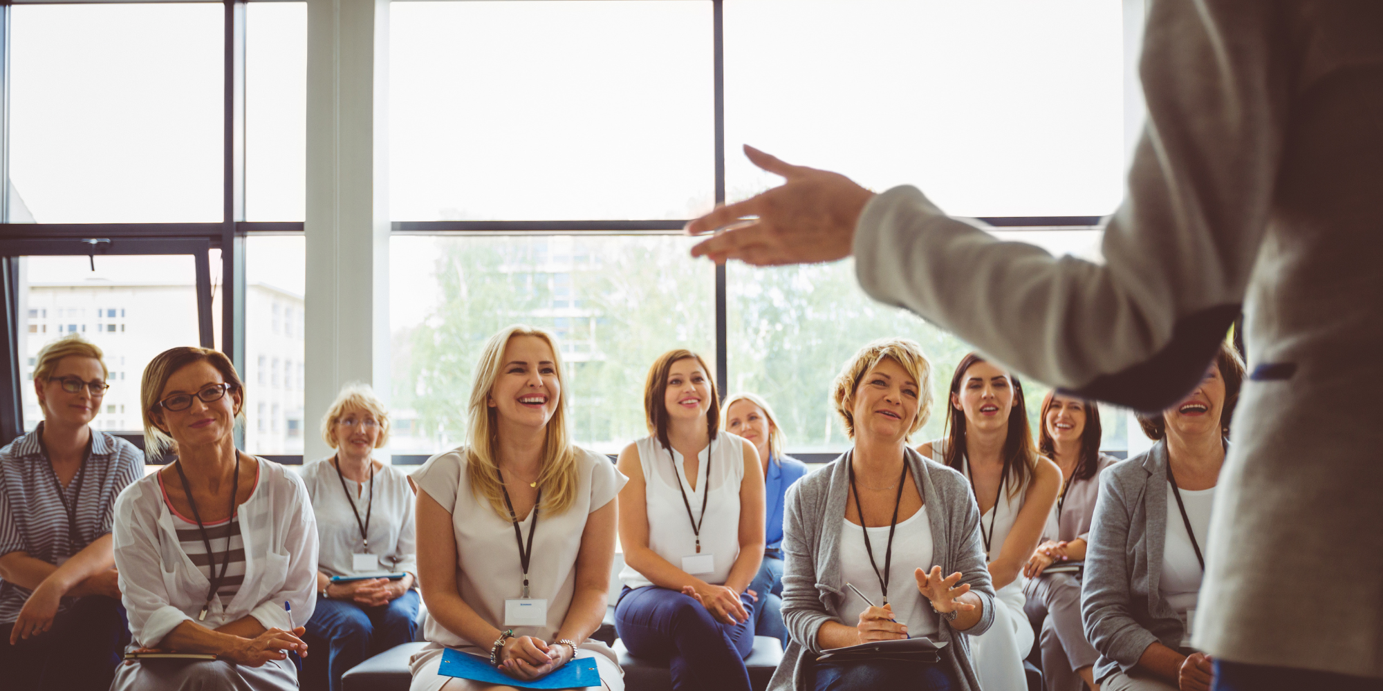 A diverse group of women attending training, listening attentively and smiling as a speaker gestures in front of them.