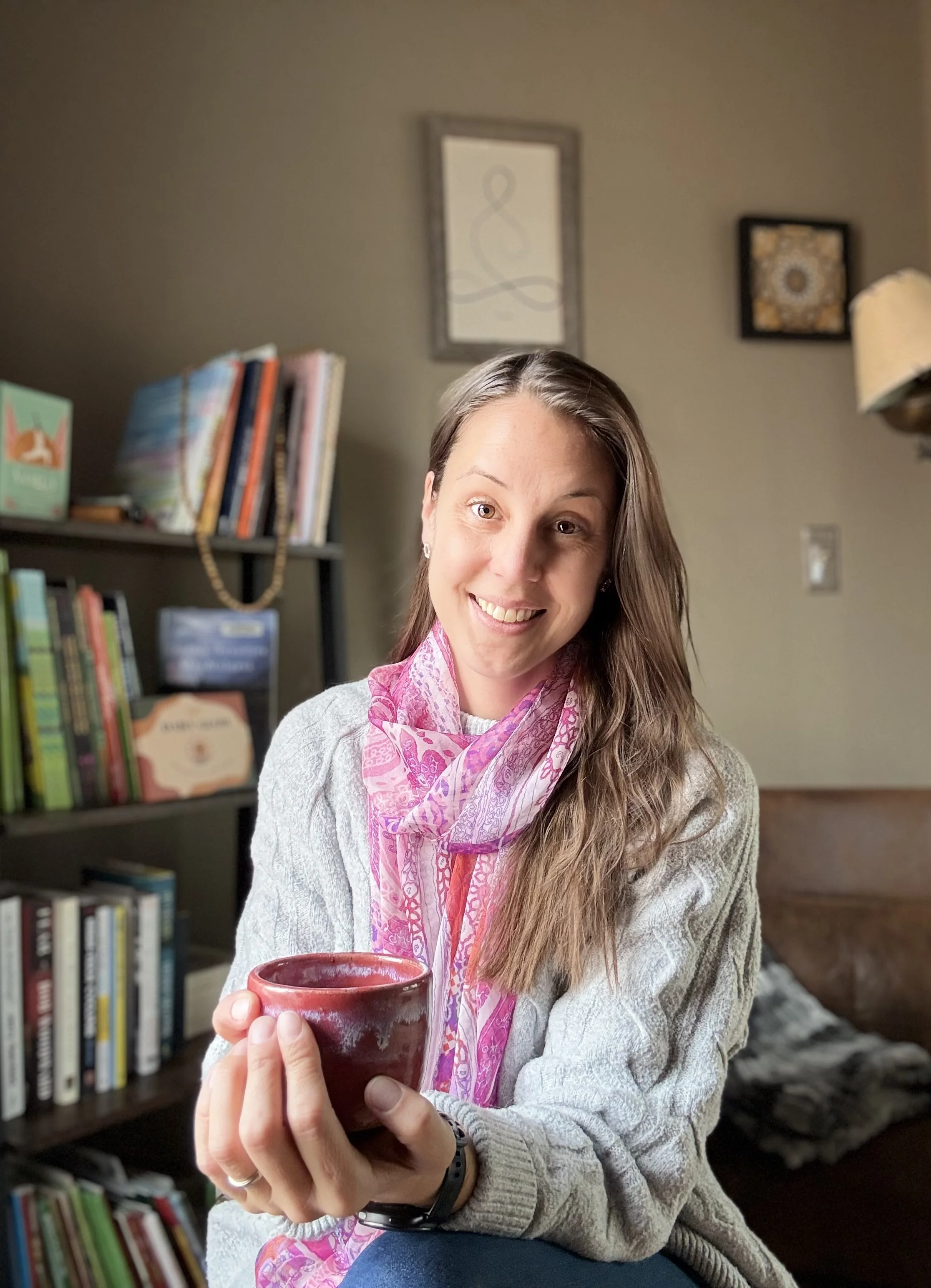 A woman with long brown hair smiling and holding a red mug in a cozy room with bookshelves and framed artwork on the wall.