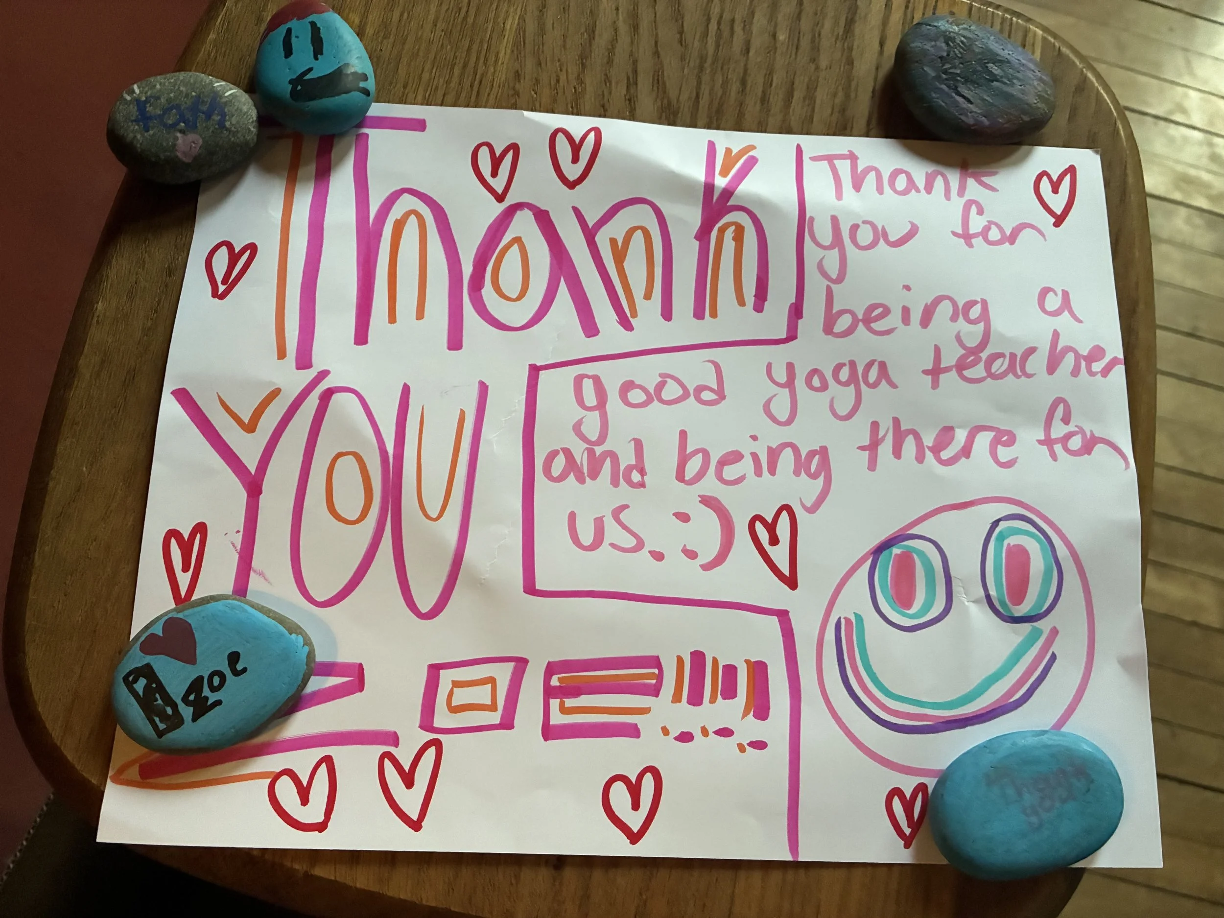 A handwritten thank you note with colorful markings and red hearts, surrounded by decorated rocks on a wooden surface.