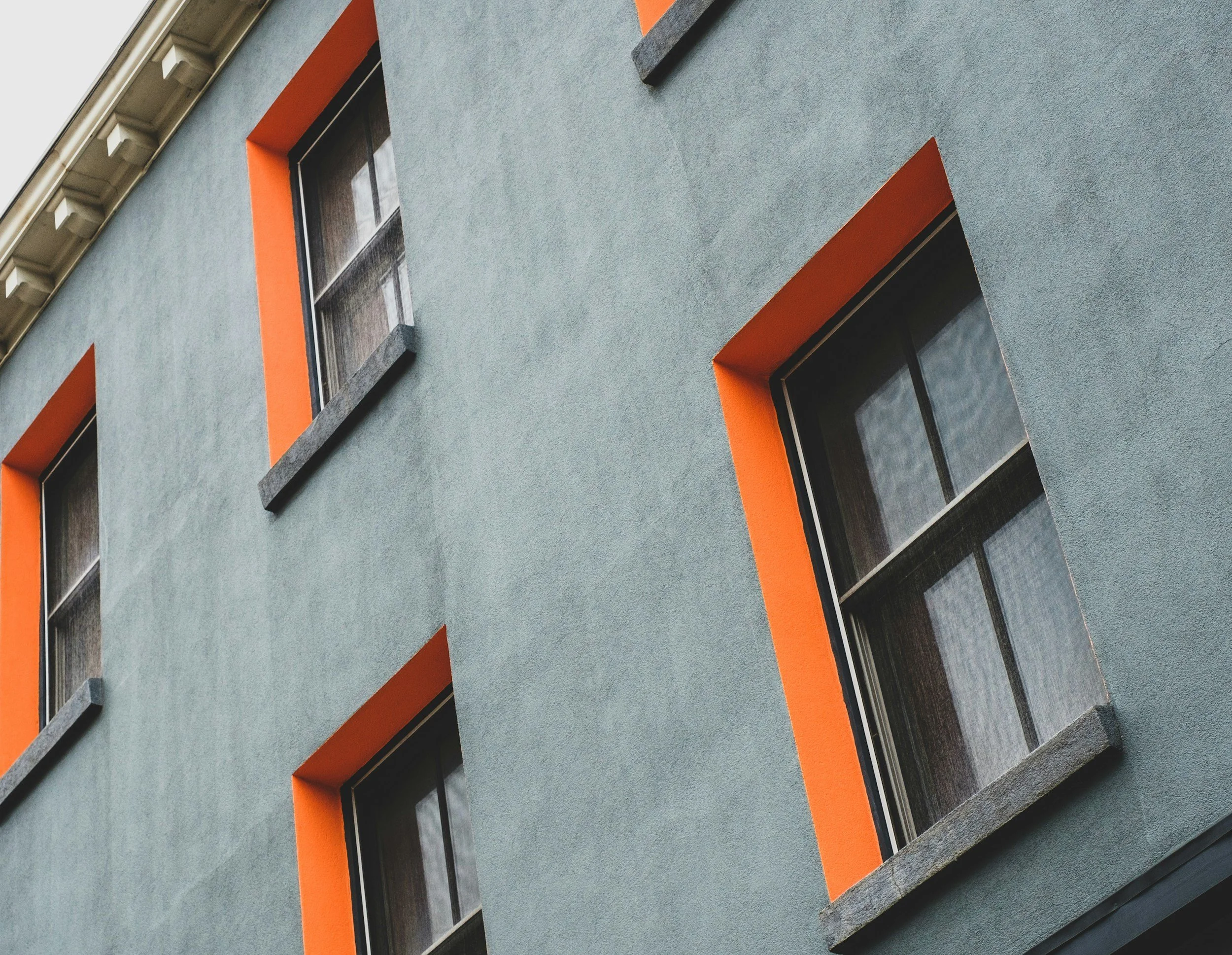 Close-up view of a modern building facade with rectangular windows, gray stone frames, orange accents around windows, and a textured blue-gray exterior wall.