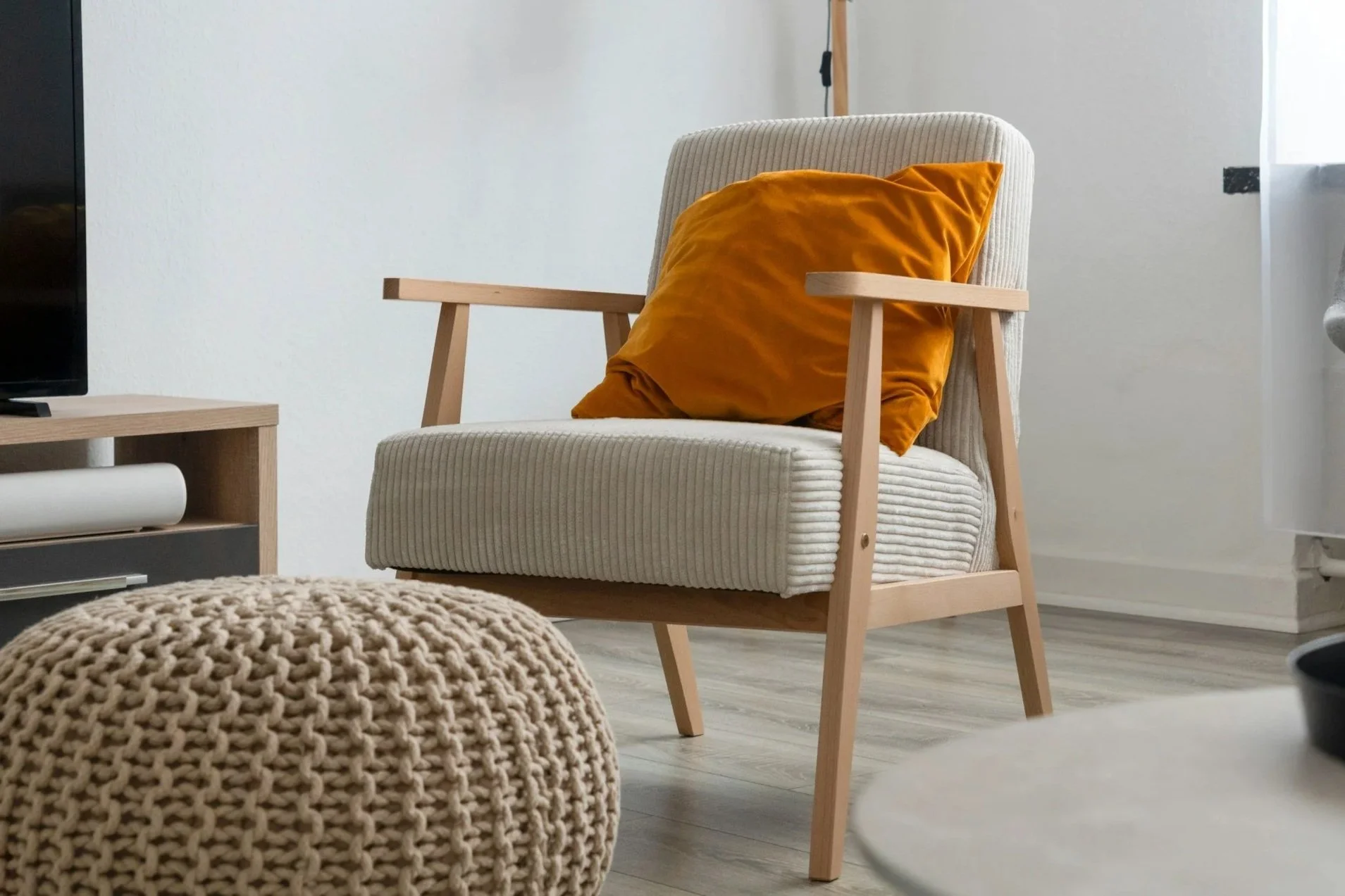 A beige armchair with a mustard-colored pillow in a modern living room, with a wooden side table and a textured beige pouf in the foreground.