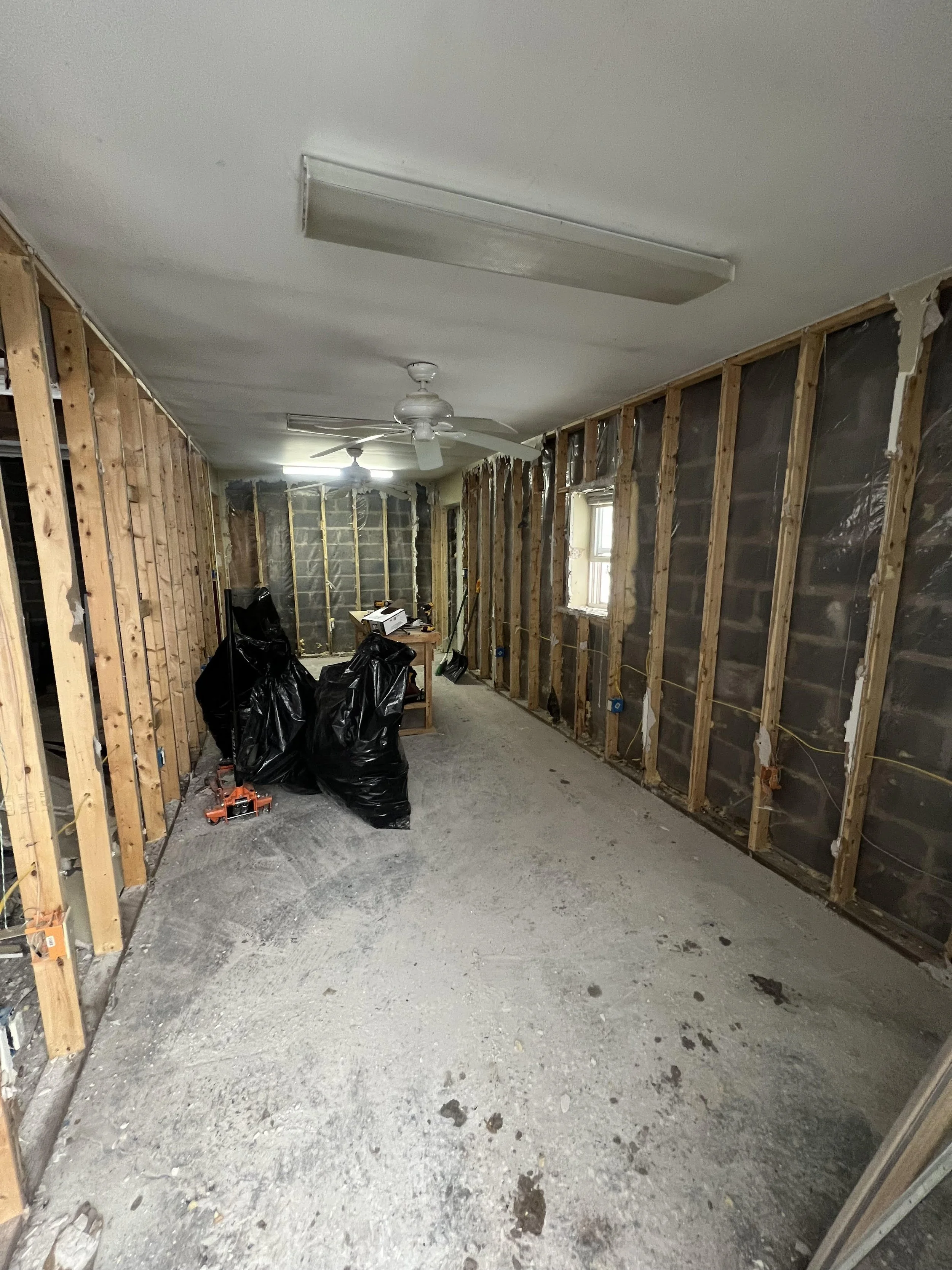 A room under renovation with exposed wall studs and plastic covering on the walls, two ceiling fans, a fluorescent light fixture, and construction materials and tools on the floor.