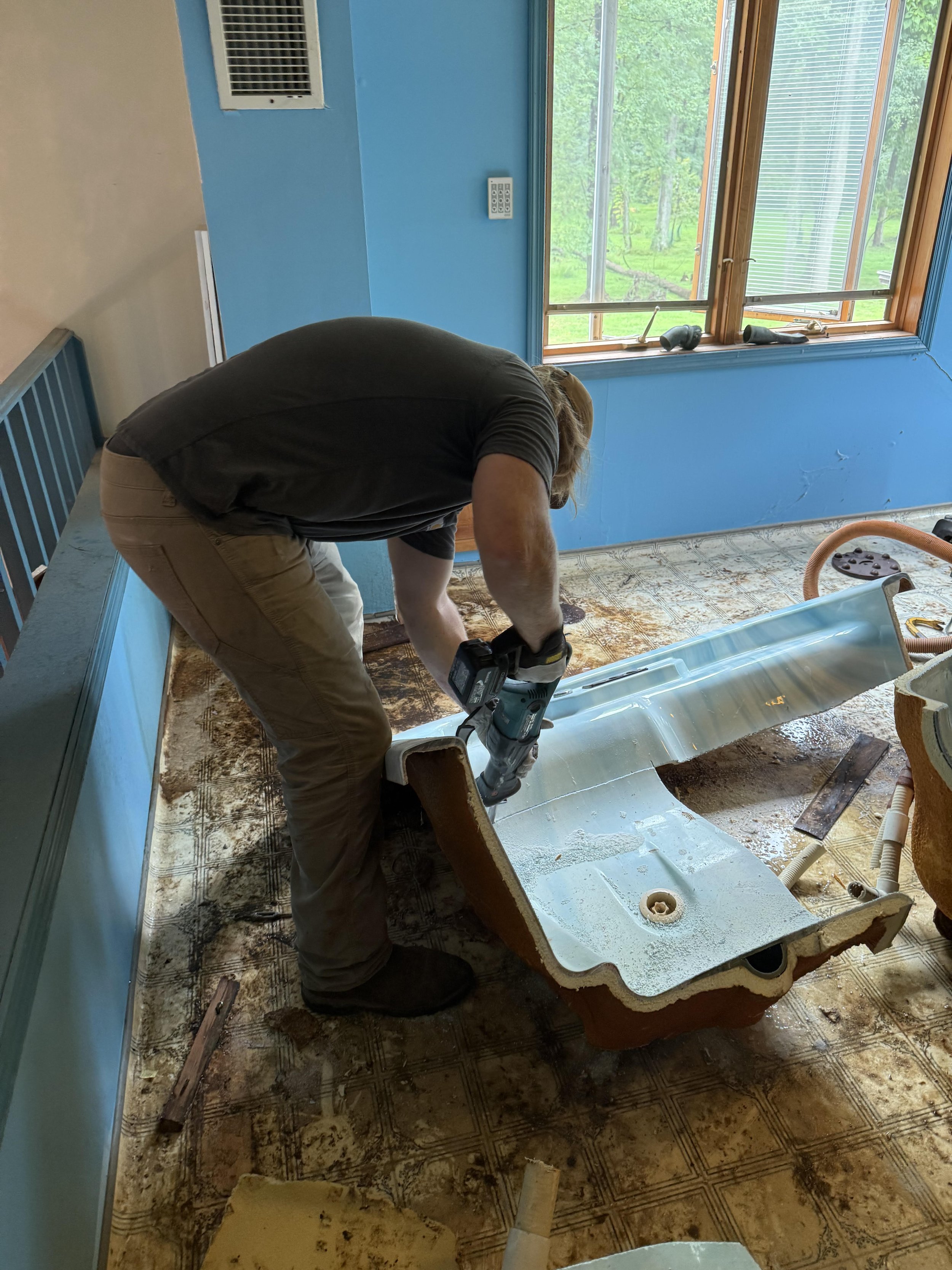 A person bending over and working on a bathroom sink with a power tool. The room has blue and beige walls, a window with green trees outside, and a patterned floor. The sink is detached and lying on the floor, with pipes and tools around.