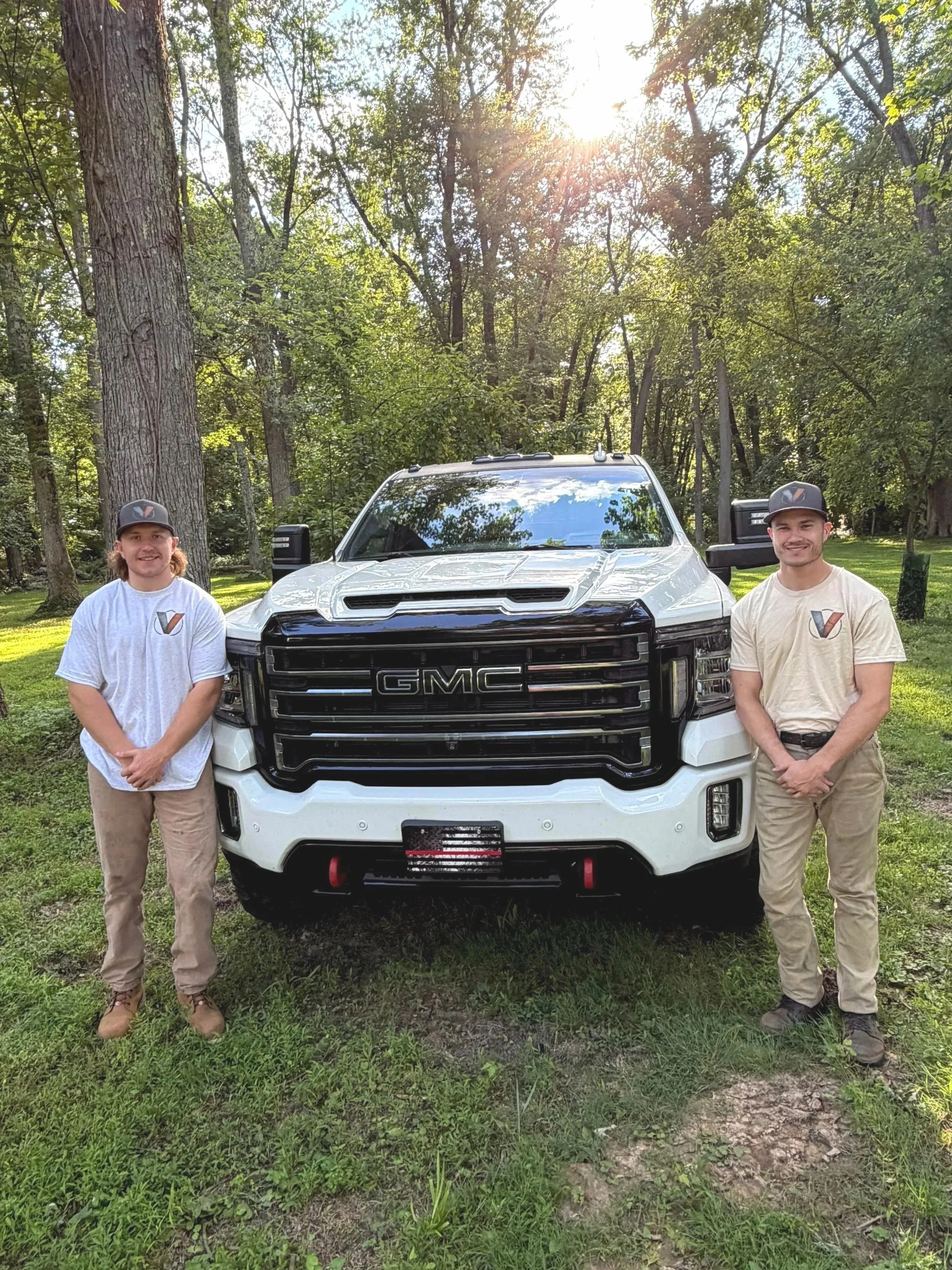 Two young men standing on either side of a white GMC truck outdoors in a wooded area on a sunny day.
