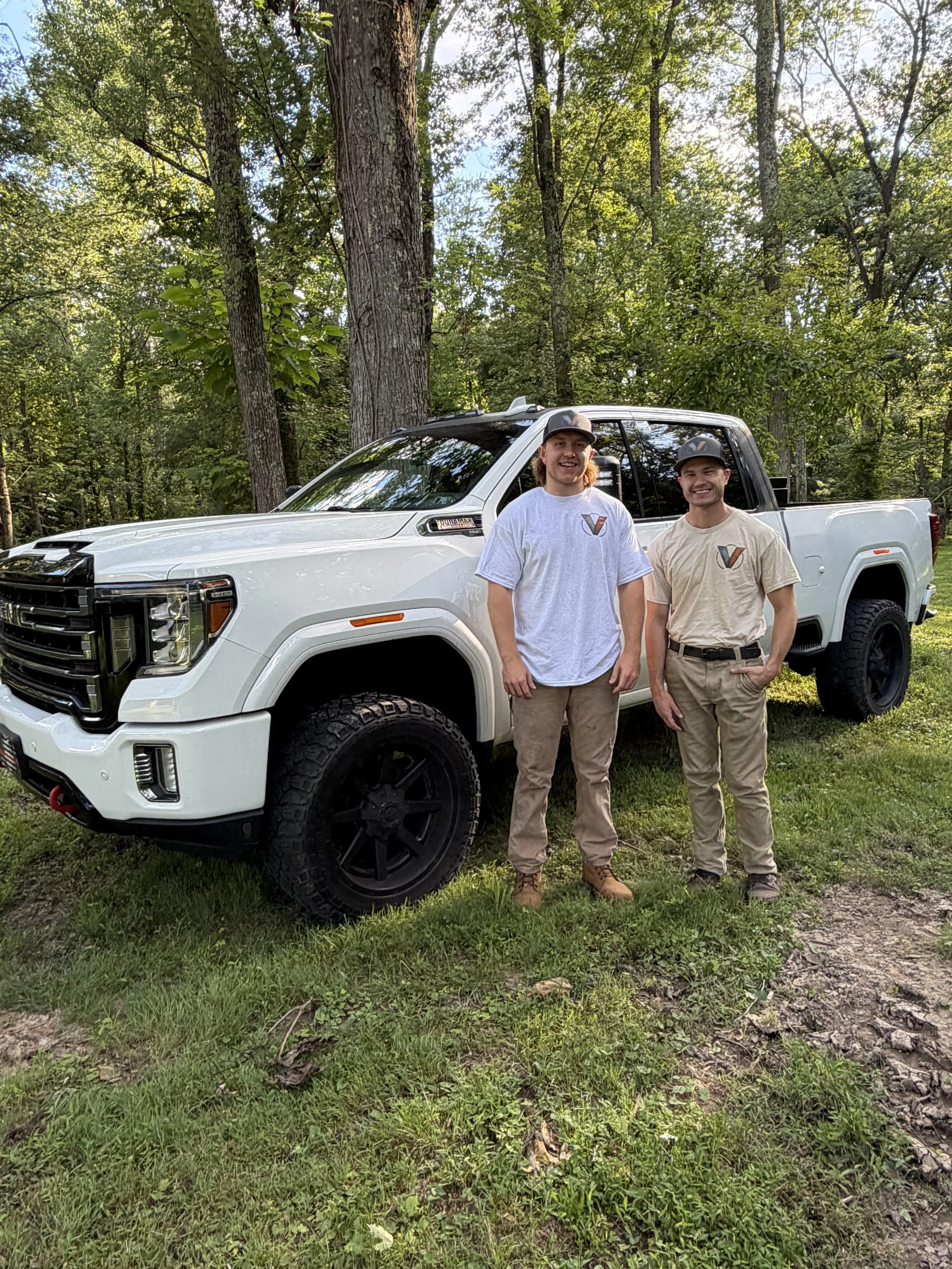 Two men stand outdoors in front of a white pickup truck in a forested area with tall trees and green foliage.
