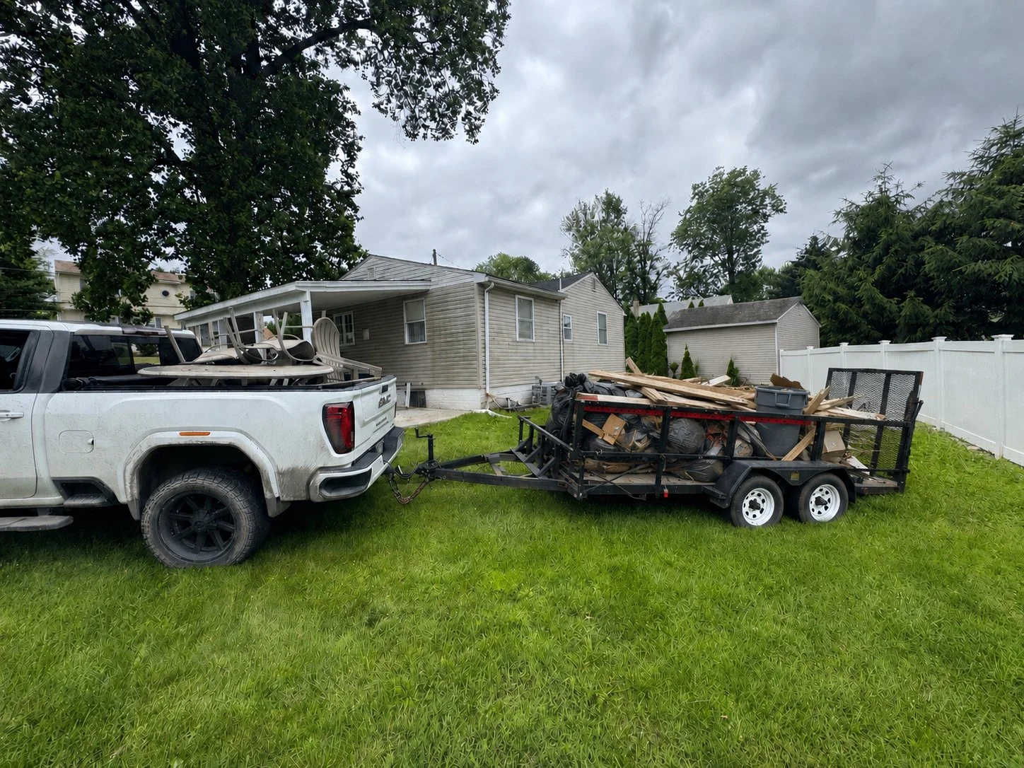 A white pickup truck with a trailer attached, filled with construction debris and wooden planks, parked on a grassy backyard near houses and trees under a cloudy sky.