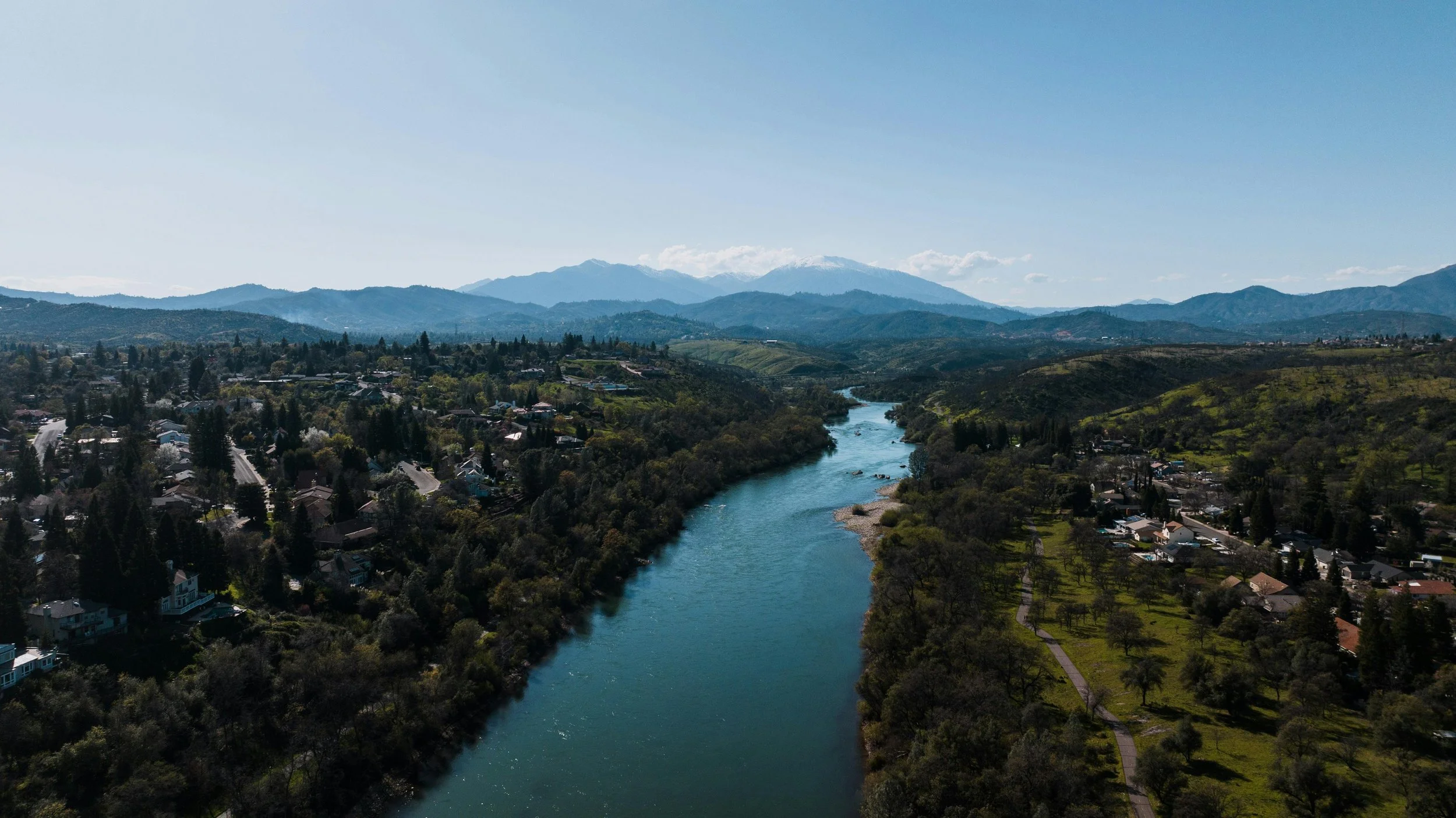 Aerial view of a river flowing through a lush green valley with houses, trees, and mountains in the background under a clear blue sky.