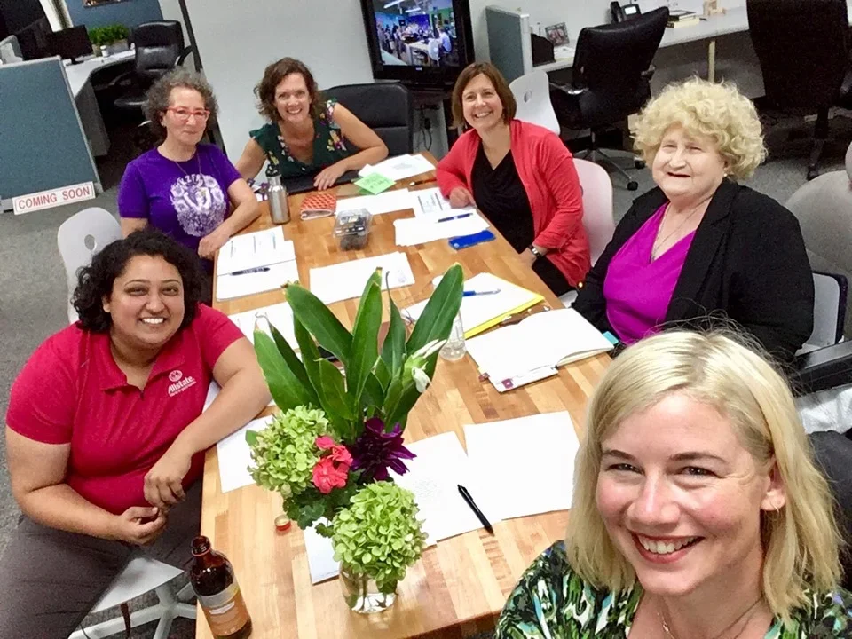 Six women sitting around a conference table with notebooks and pens, smiling, with a large floral arrangement in the center. Office environment with cubicles and monitors in the background.