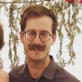 A man with brown hair, glasses, and a mustache smiles outdoors with greenery in the background.