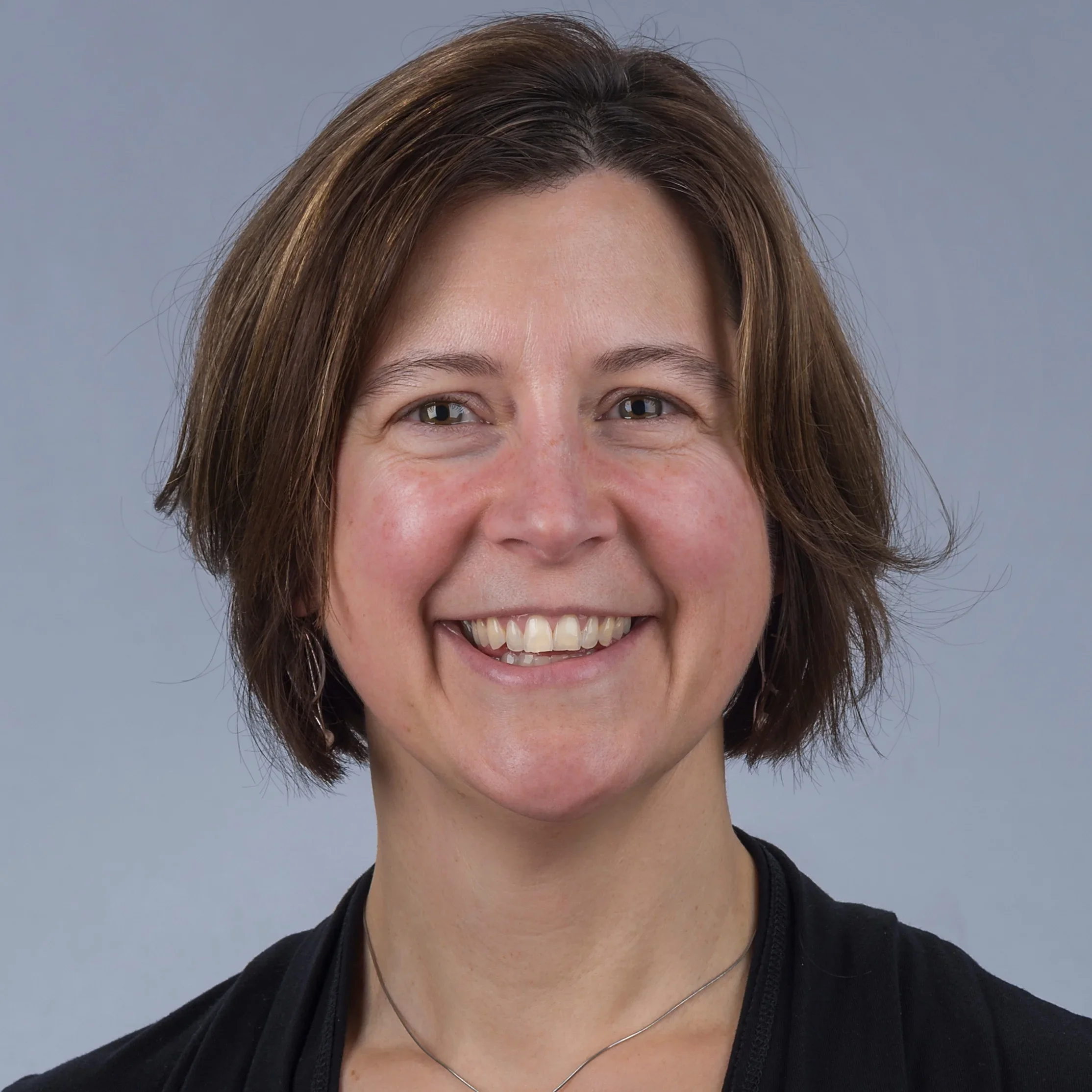 Close-up of a woman with short brown hair, smiling, wearing a black top and a silver necklace, against a gray background.