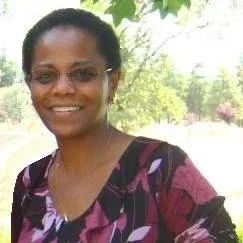 A woman with glasses, smiling outdoors in a park or garden with green trees in the background.