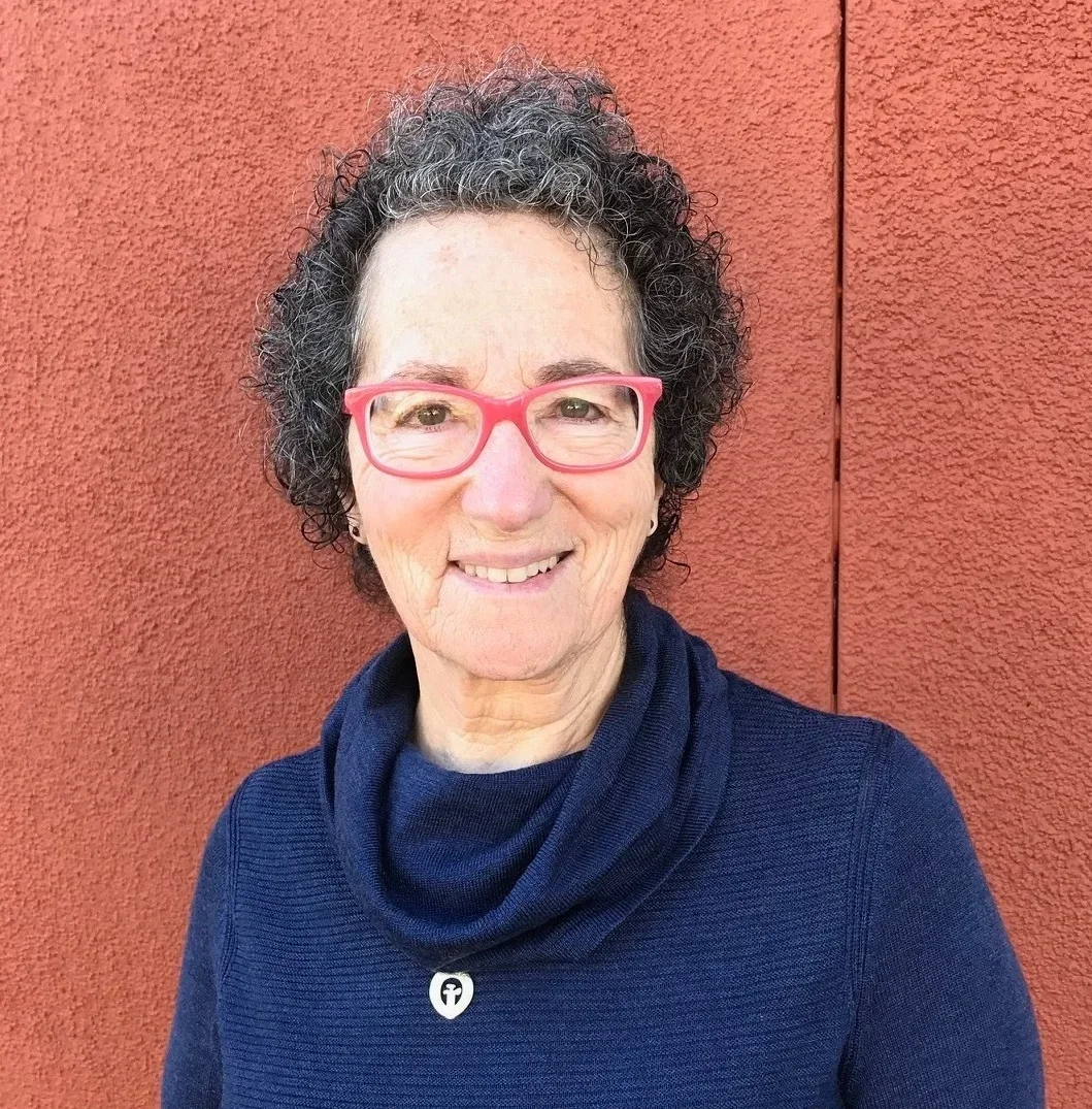 A smiling woman with curly gray hair and pink glasses, wearing a dark blue top with a cowl neck, standing in front of a red textured wall.