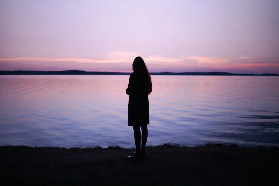 Silhouette of a person standing by a body of water during sunset or dawn, with a purple and pink sky.