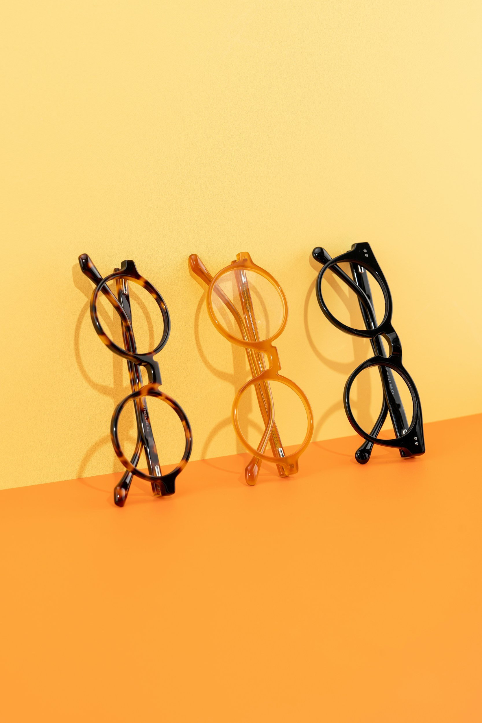 Three pairs of eyeglasses on an orange and yellow background, with the glasses frames in tortoiseshell, orange, and black.