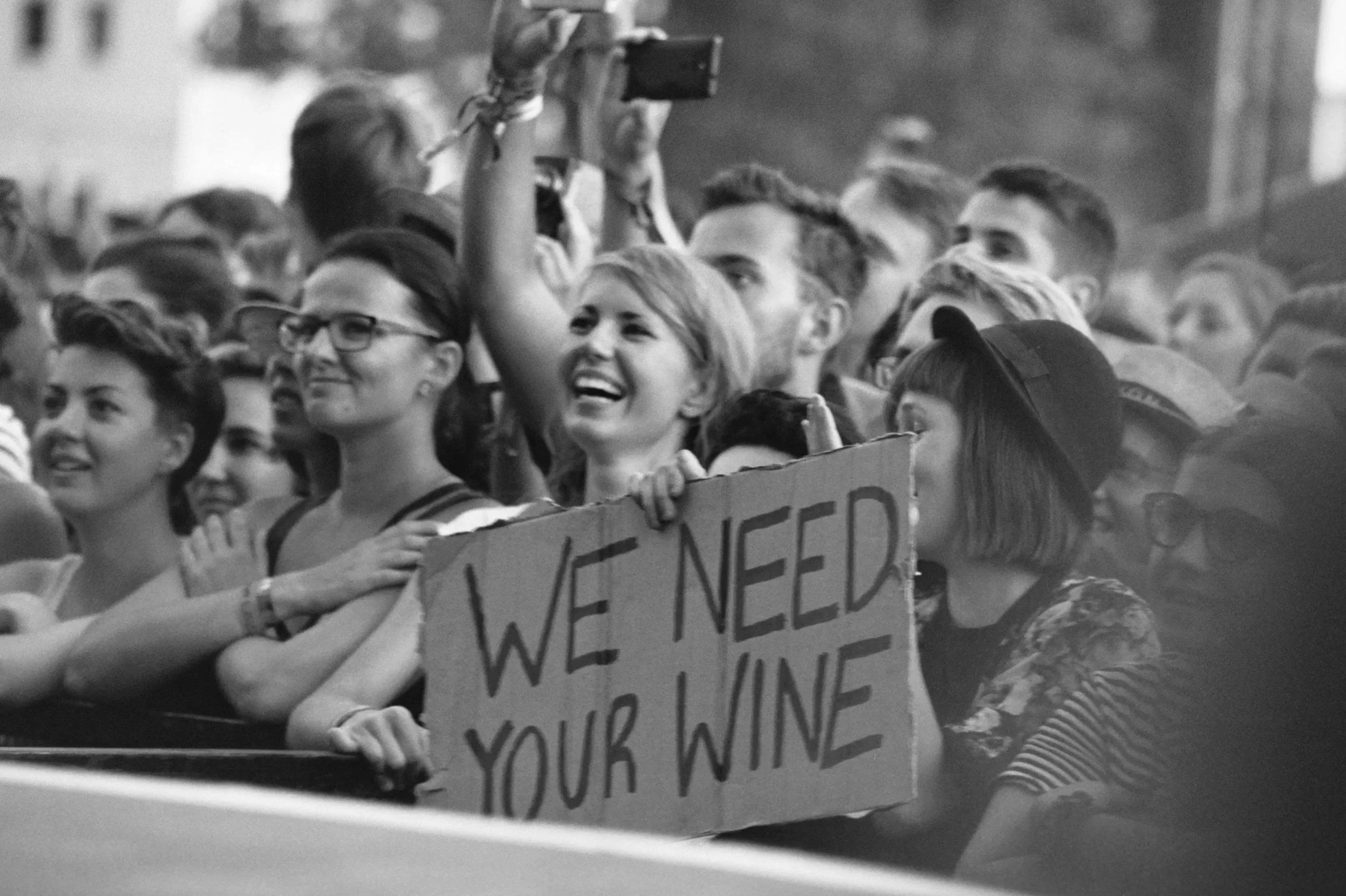 A group of people cheering with a sign that reads "We Need your Wine".
