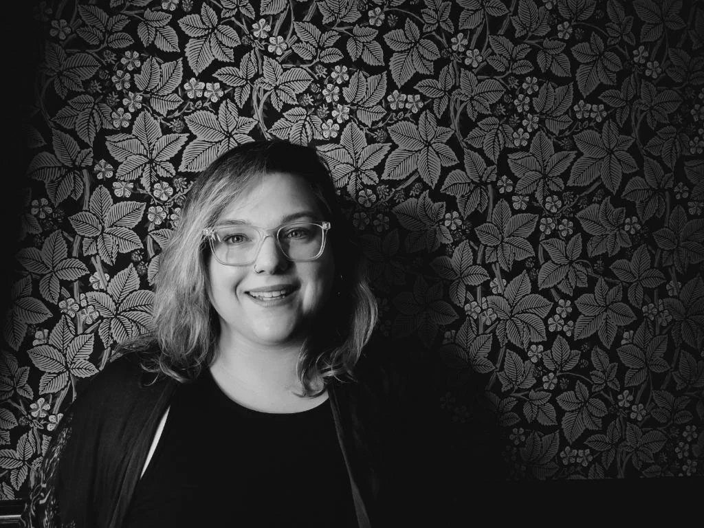 Black and white portrait of a smiling young woman with glasses, shoulder-length hair, standing in front of a decorative leafy patterned background.