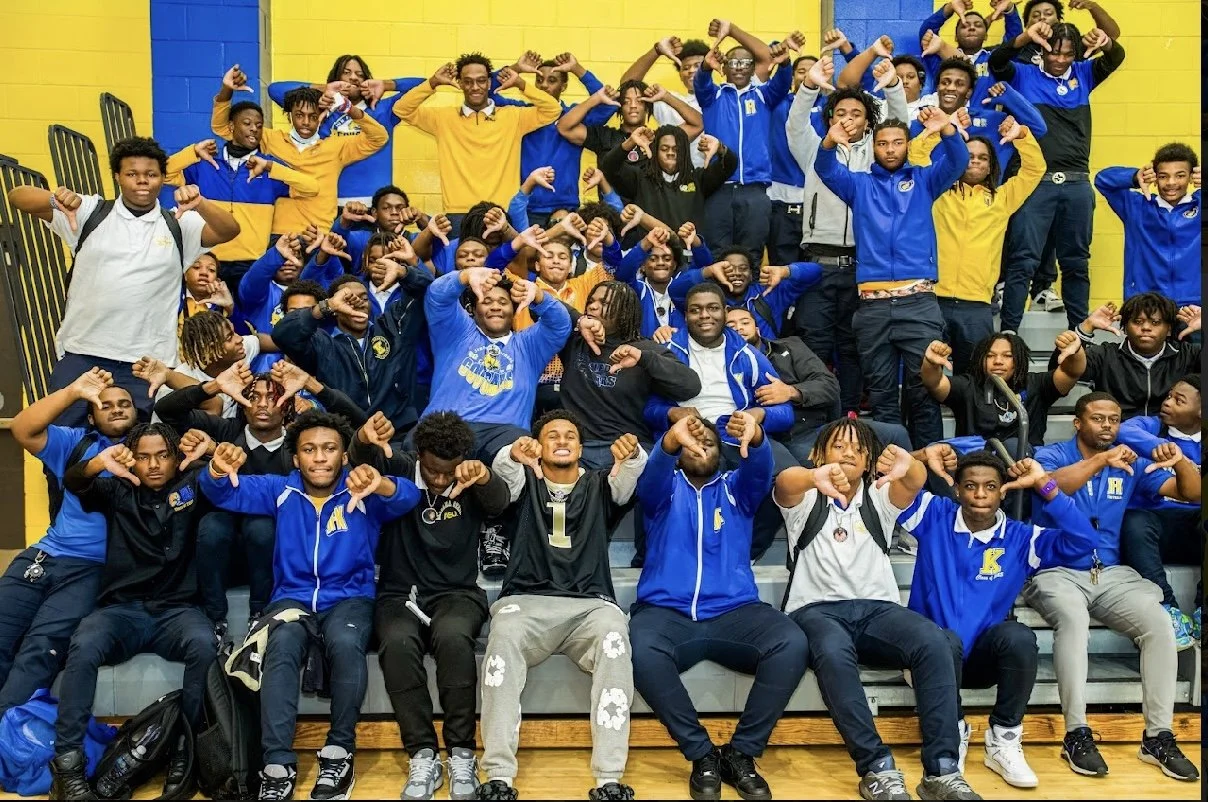 Group of students and a coach posing in a gymnasium, raising their fists, with a yellow and blue wall background.