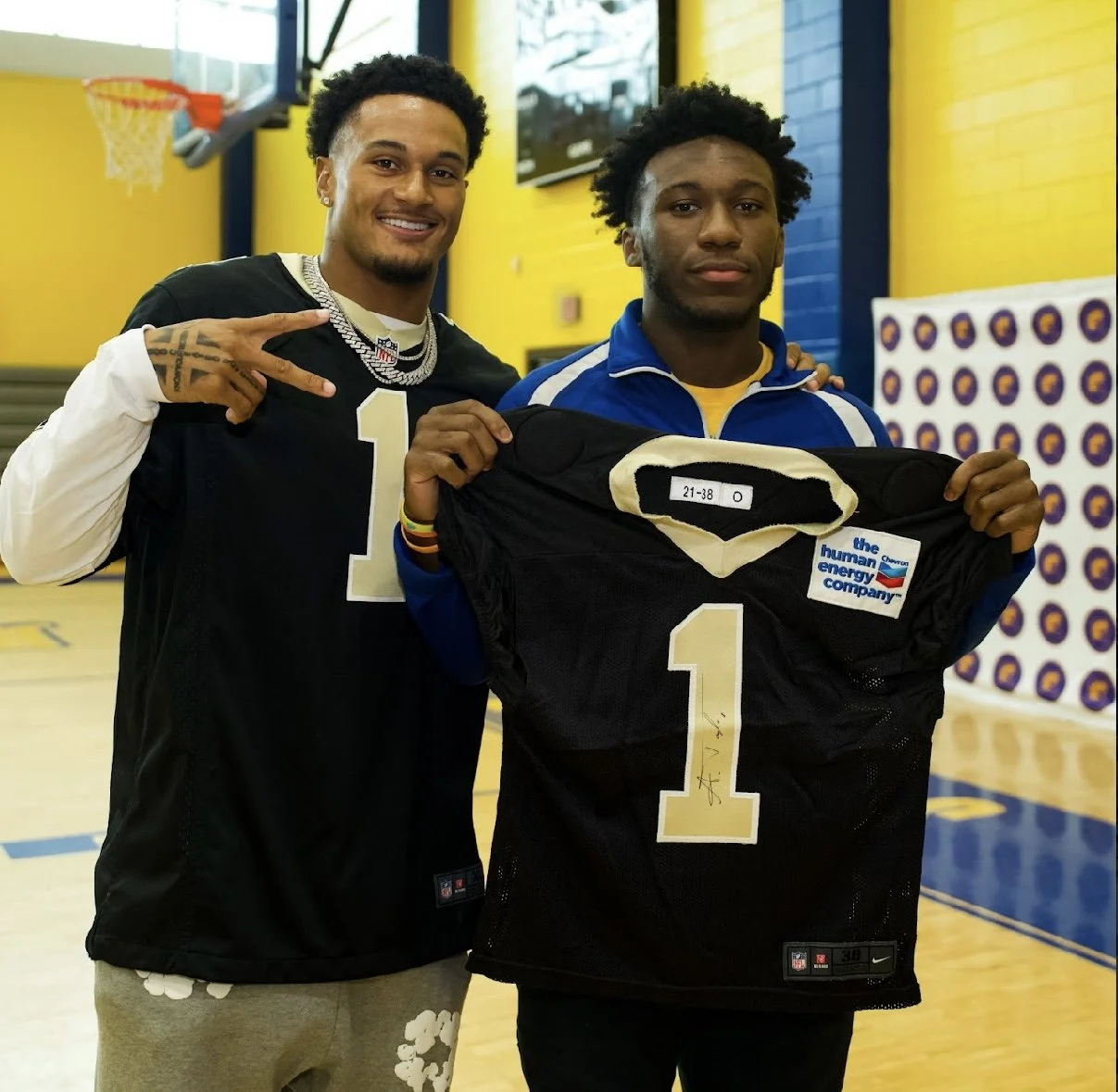 Two men standing in a gymnasium, one giving a peace sign and the other holding a jersey with number 1. The man on the left is wearing a black football jersey with white long sleeves, a silver chain, and multiple bracelets. The man on the right is in 