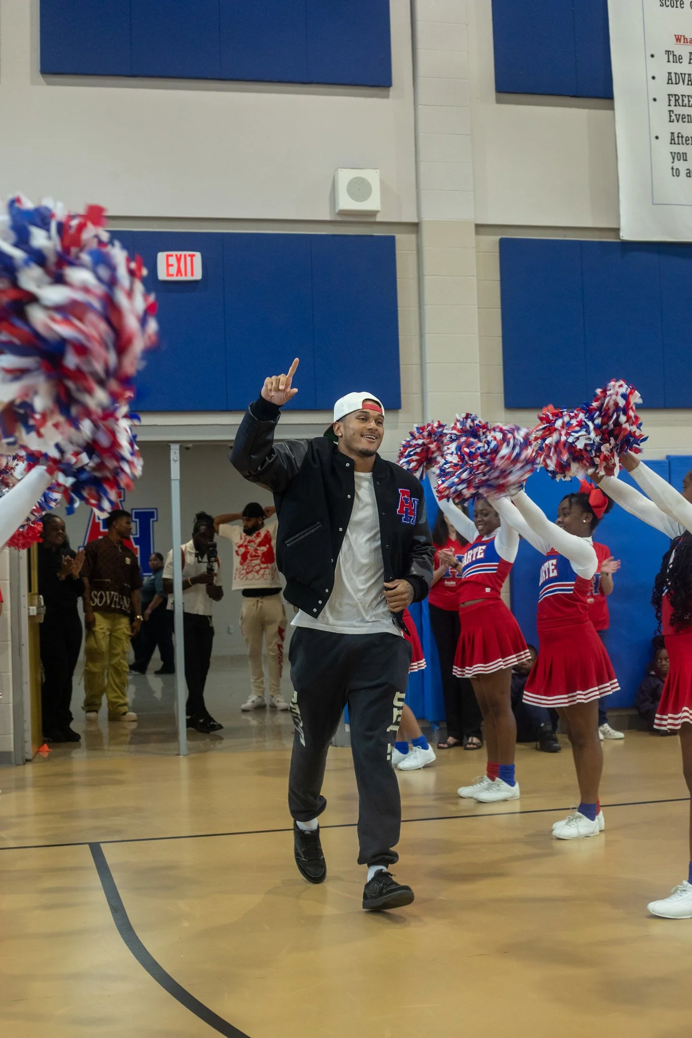 A young man in a white hat and black jacket dancing at a school gym, surrounded by cheerleaders in red and blue uniforms holding pom-poms, with spectators in the background.