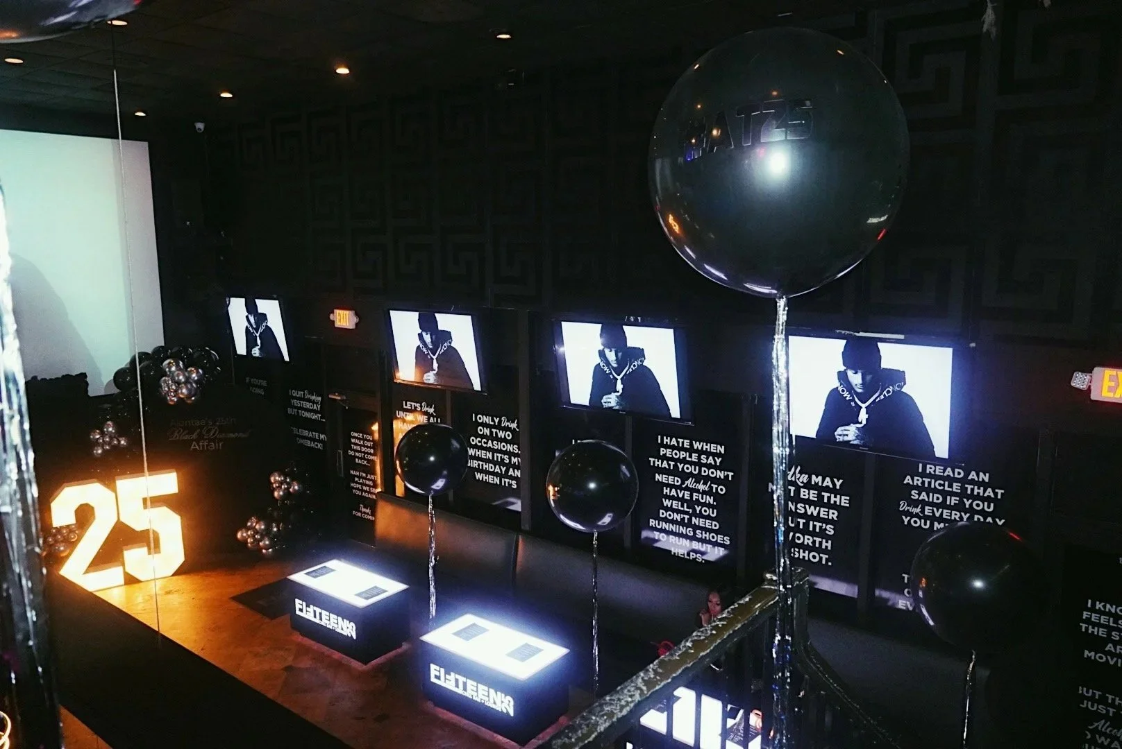 An indoor nightclub or lounge decorated for a 25th celebration with black balloons, illuminated number 25, multiple screens showing a man, and various posters with quotes, with a dark interior and subdued lighting.