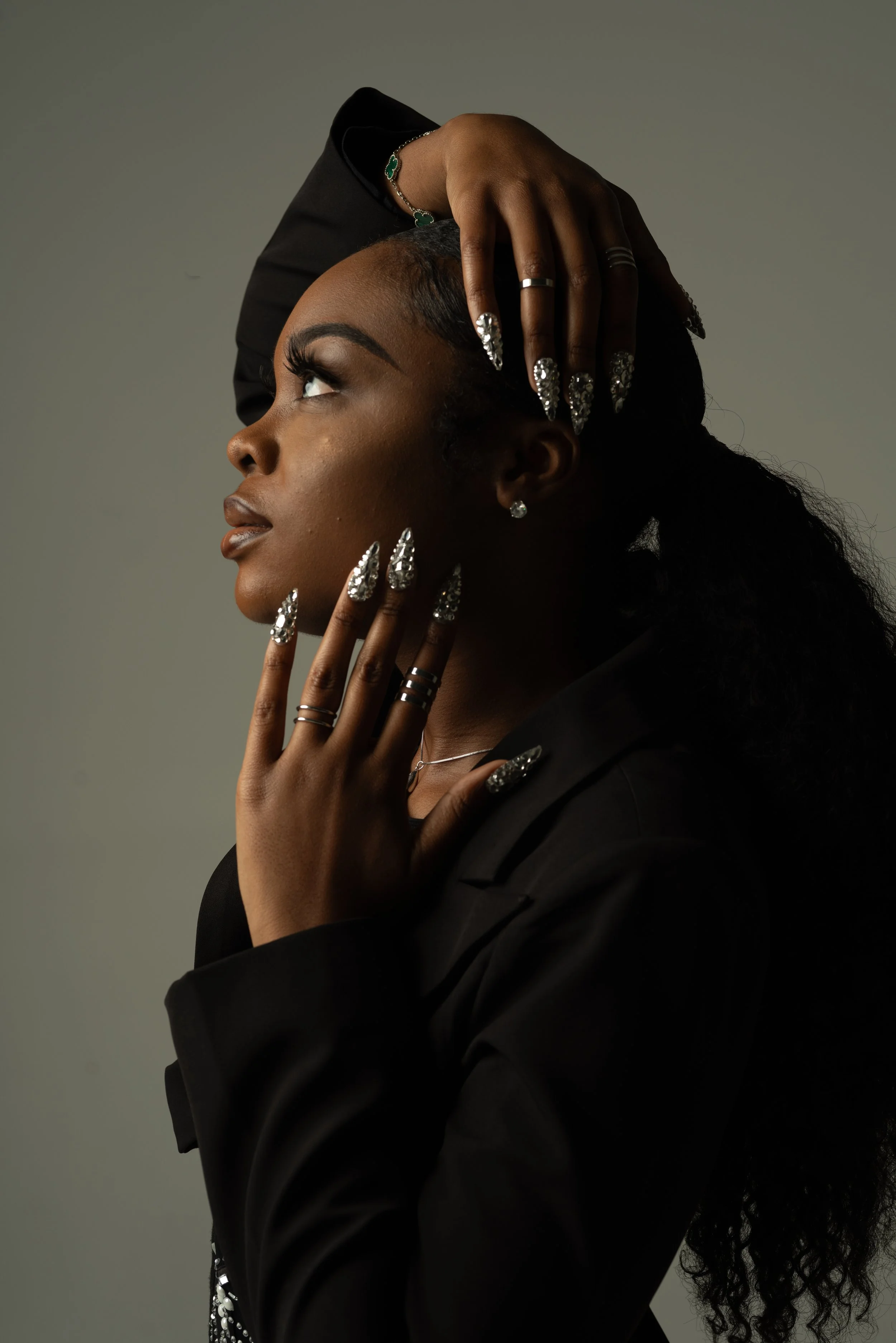 A woman with dark skin and long, curly hair is posing with her head tilted back and her hand resting on her forehead. She is wearing multiple rings, a bracelet, earrings, and a necklace, and has long, decorated nails. She is dressed in a black outfit against a plain, neutral background.