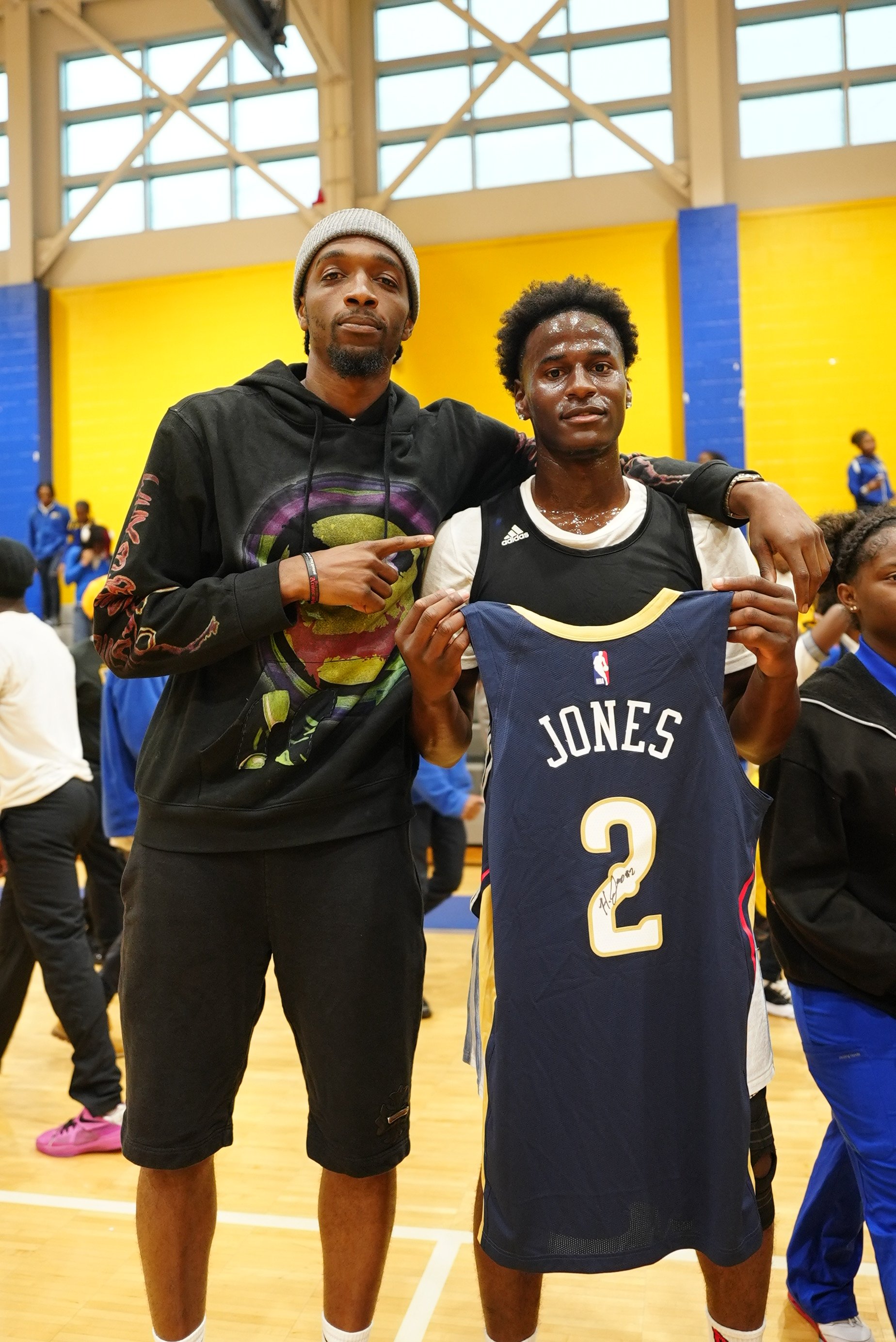 Two young men standing inside a gymnasium, one wearing a beanie and black hoodie and the other in a black and white basketball uniform, holding a jersey with the name 'Jones' and the number 2, with other people in the background.
